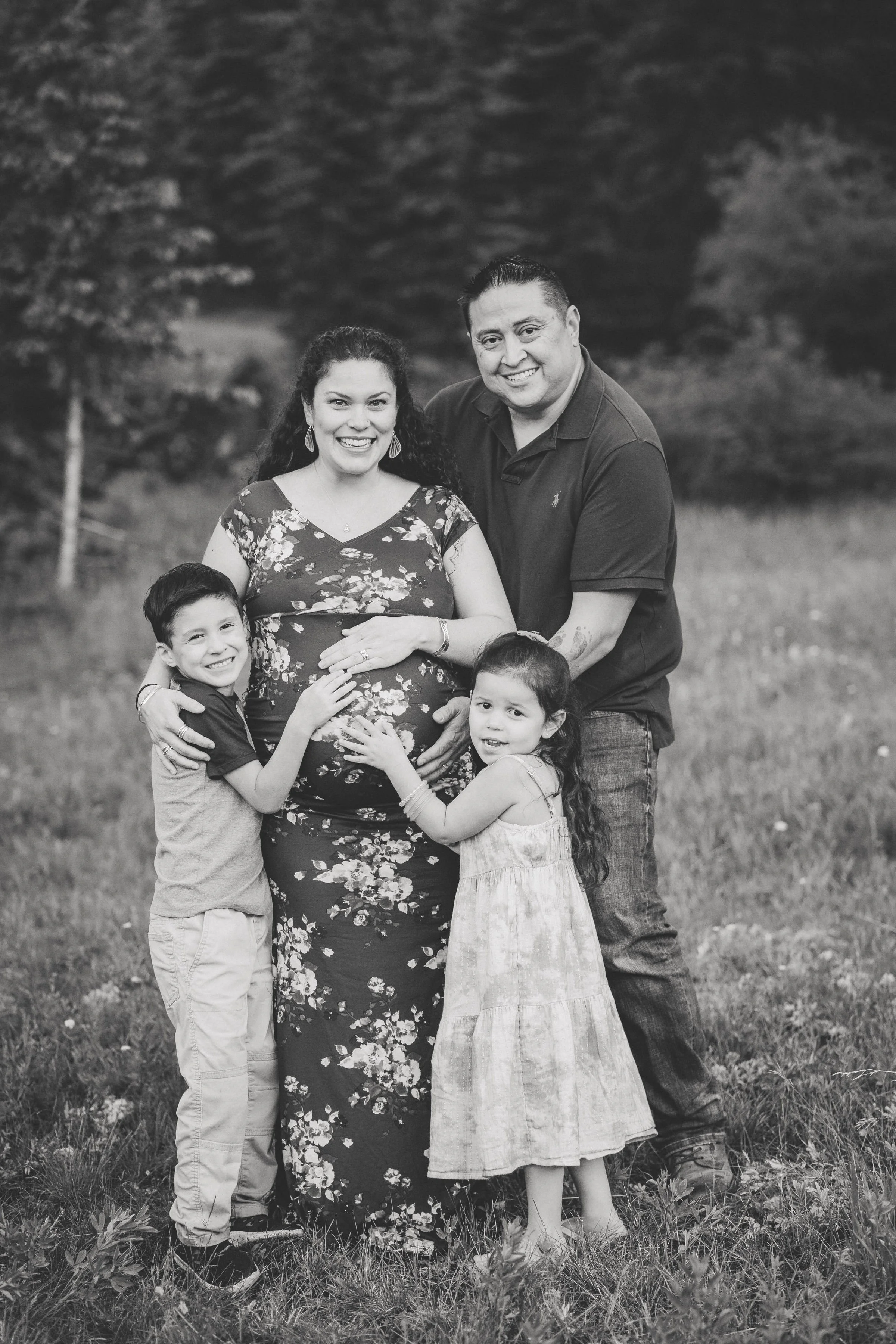 A black and white photo of a family of five standing outdoors; a pregnant woman wearing a floral dress, a man in a dark polo shirt, a young boy in a short-sleeved shirt and pants, a young girl in a light-colored dress, all smiling and embracing each 