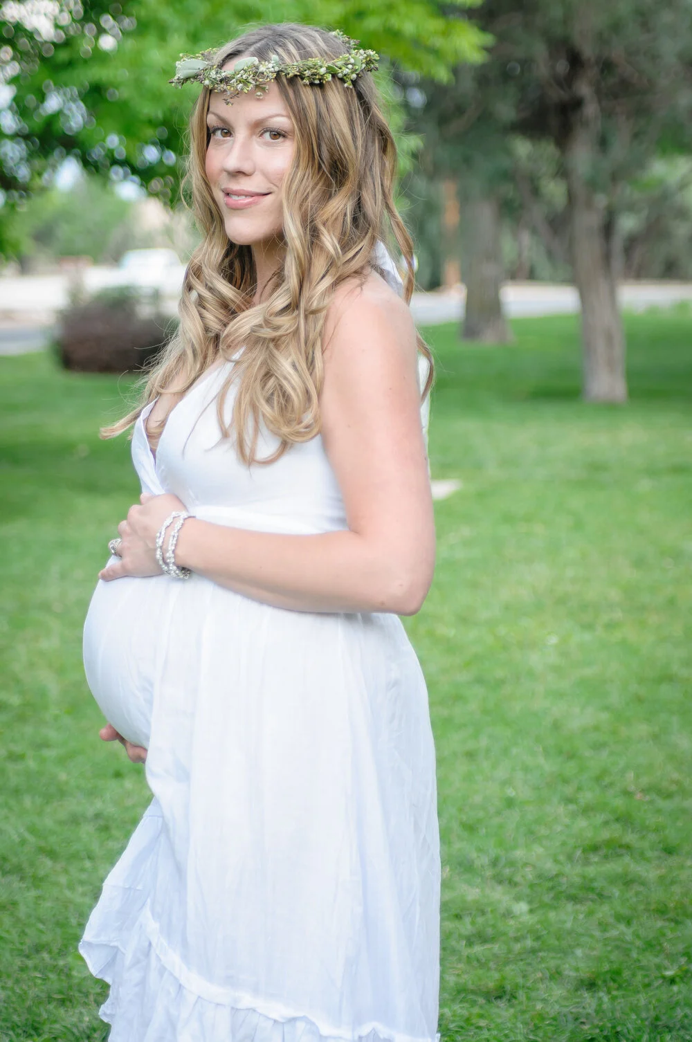 A pregnant woman with long wavy blonde hair, wearing a white sleeveless dress and a floral crown, standing outdoors on a grassy area with trees in the background, smiling and gently holding her belly.