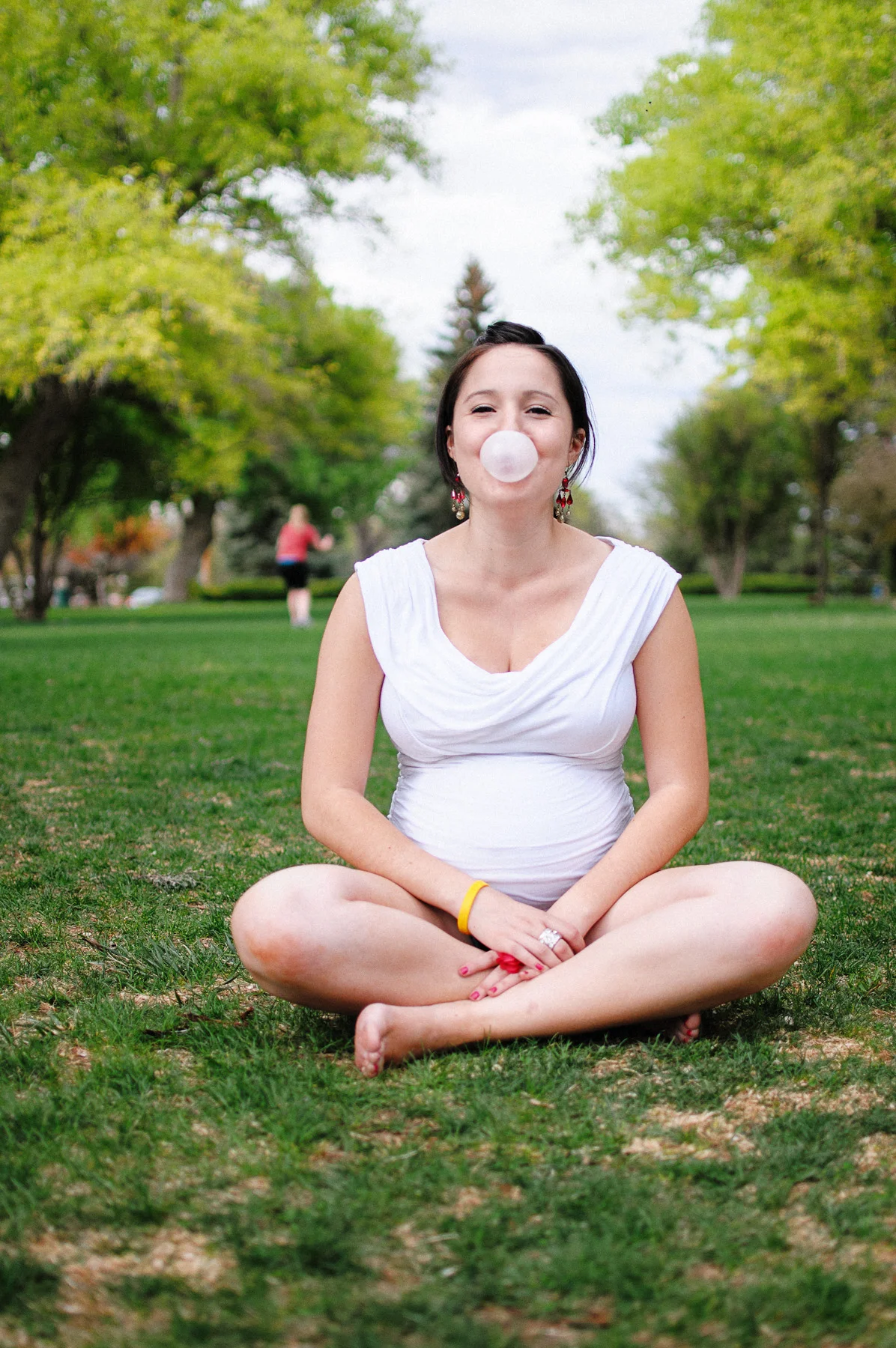 A woman sitting cross-legged on grass in a park, blowing a bubble with bubblegum, wearing a white top and earrings, surrounded by green trees.