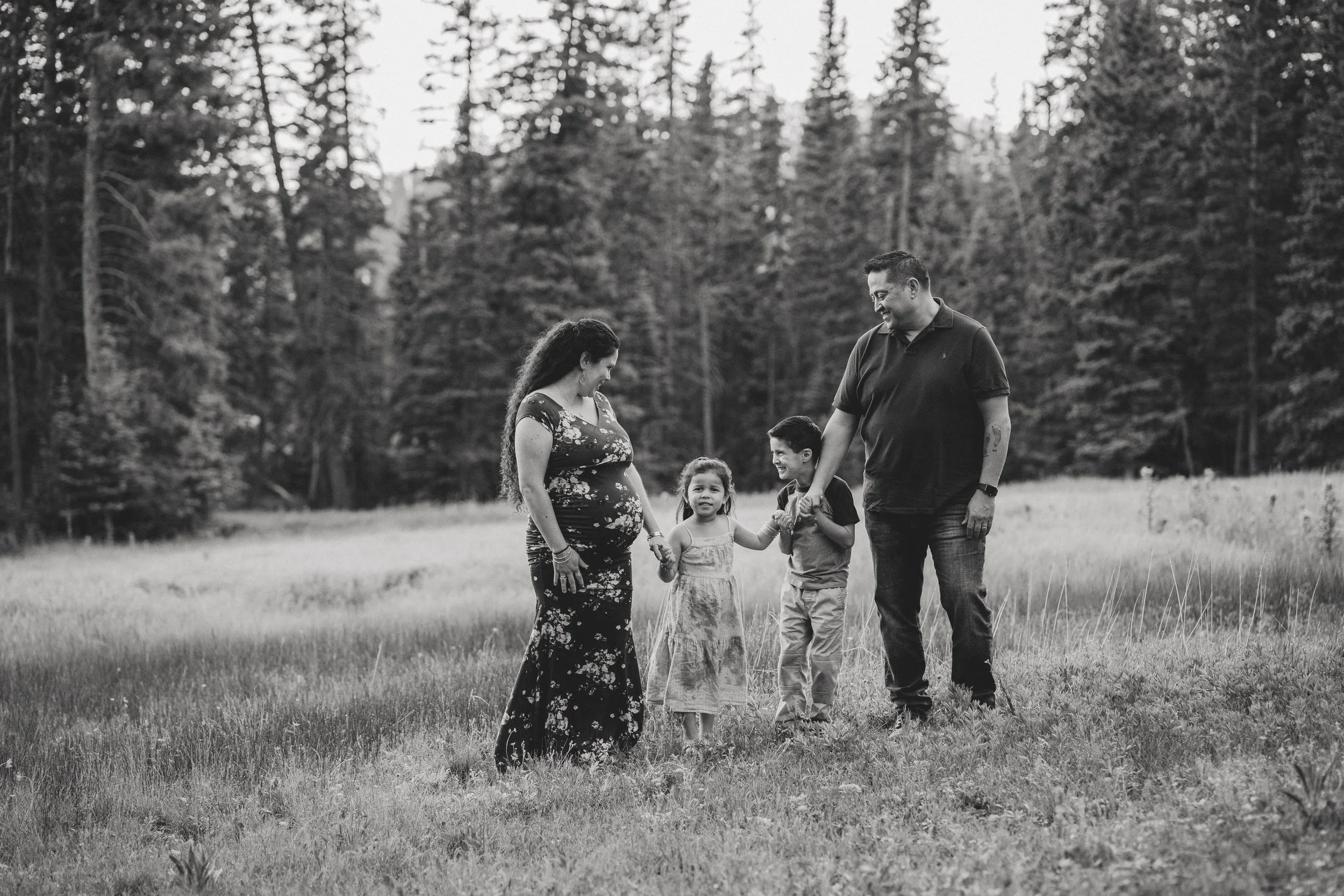 A family of five standing outdoors in a grassy field surrounded by trees, with the pregnant mother and father holding hands with two children, smiling and looking at each other in a black and white photo.