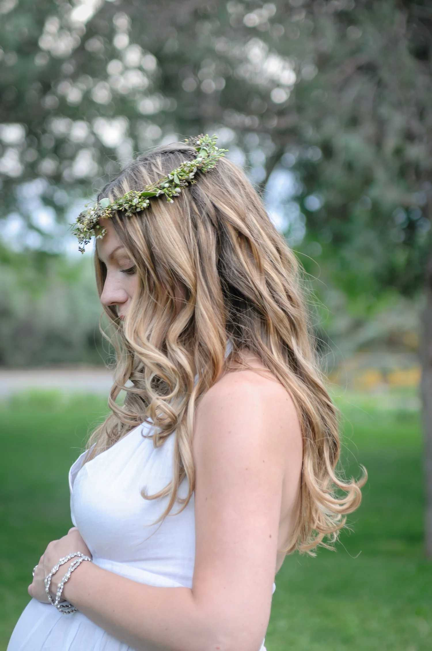 A young woman with long, wavy blonde hair wearing a flower crown made of green foliage and small white flowers, dressed in a white sleeveless dress, standing outdoors on green grass with trees in the background, looking down.