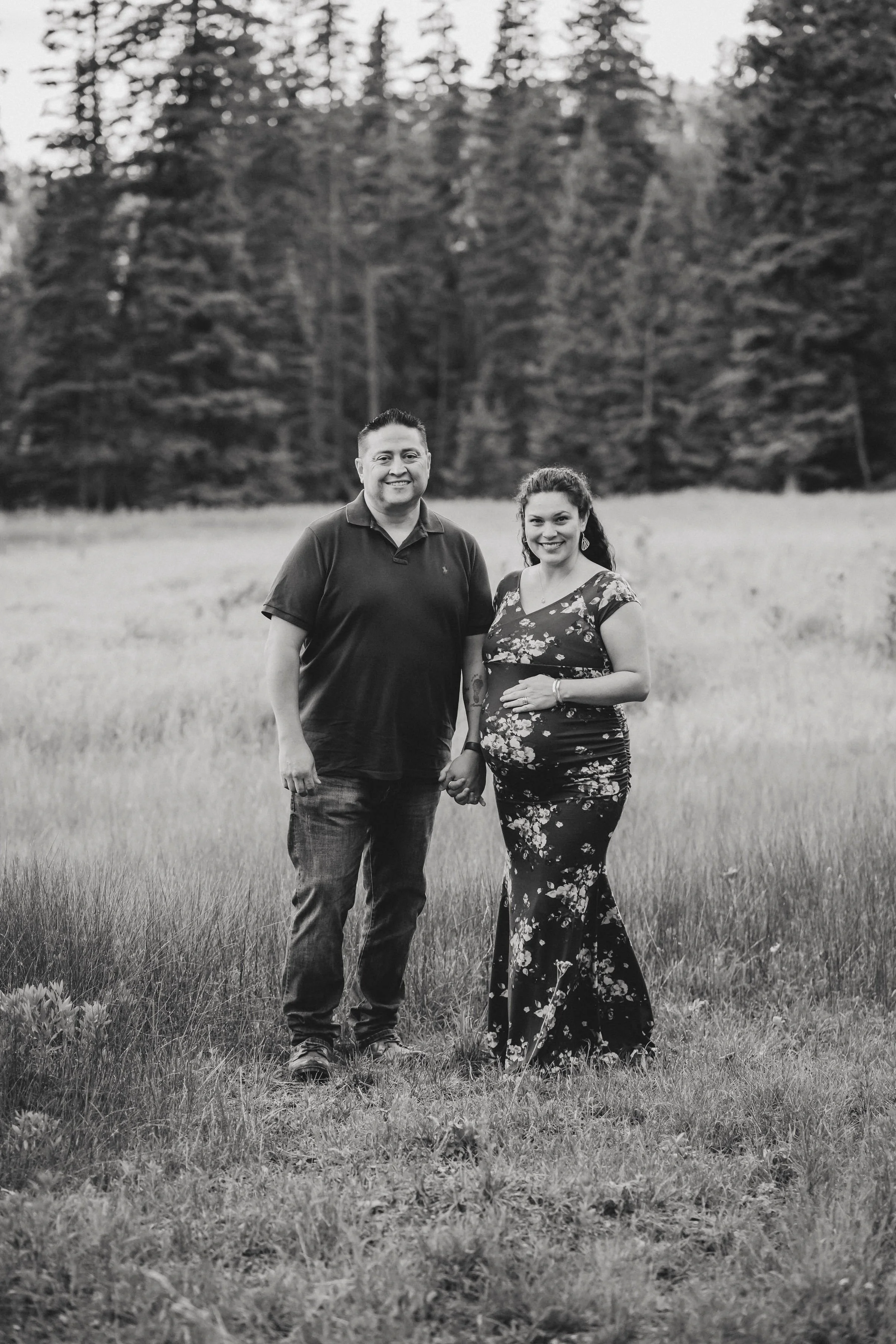 A couple holding hands in a grassy field with trees in the background, the woman is visibly pregnant, and they are both smiling.