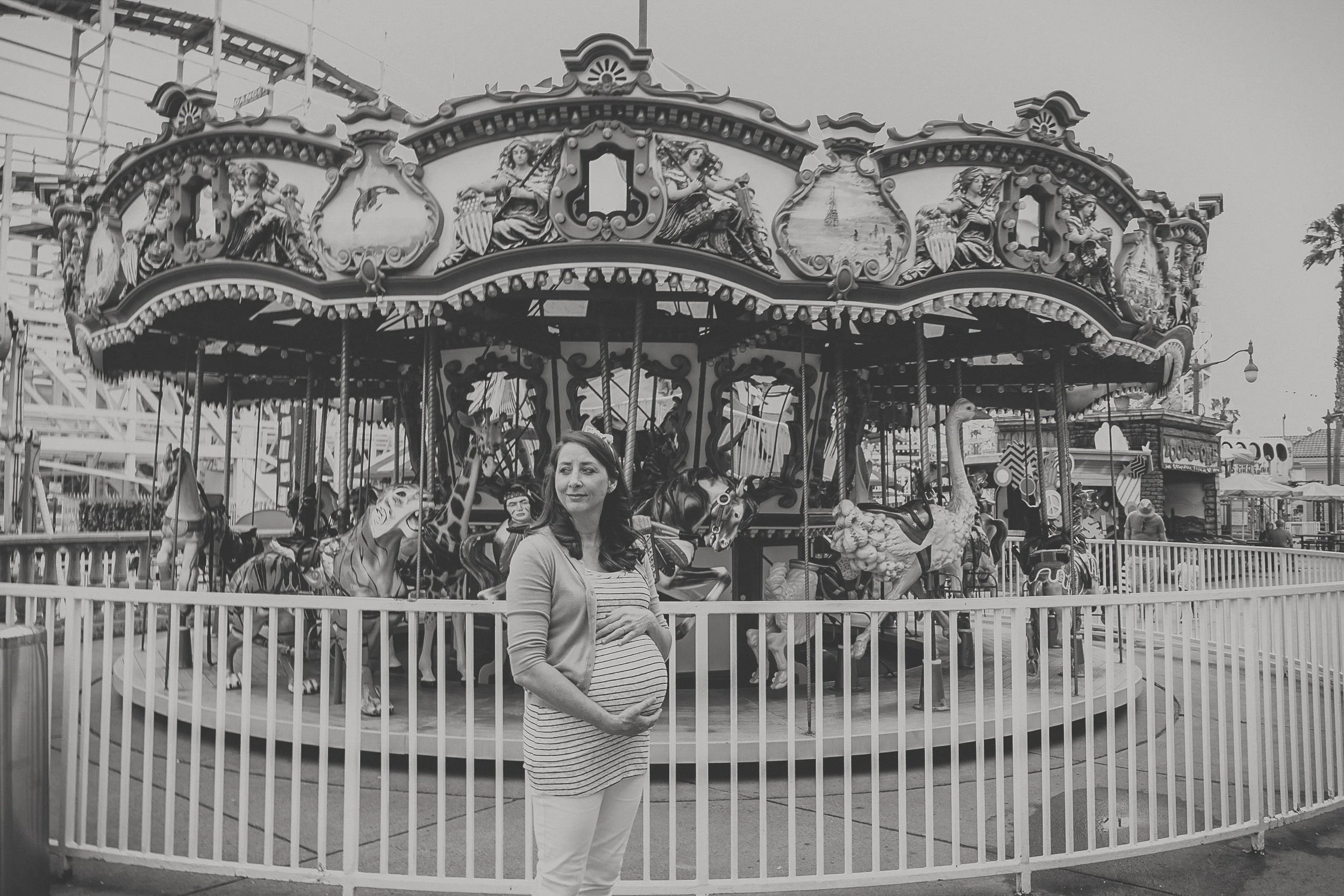 A pregnant woman standing in front of a vintage carousel at an amusement park, with her hand on her belly, in black and white.