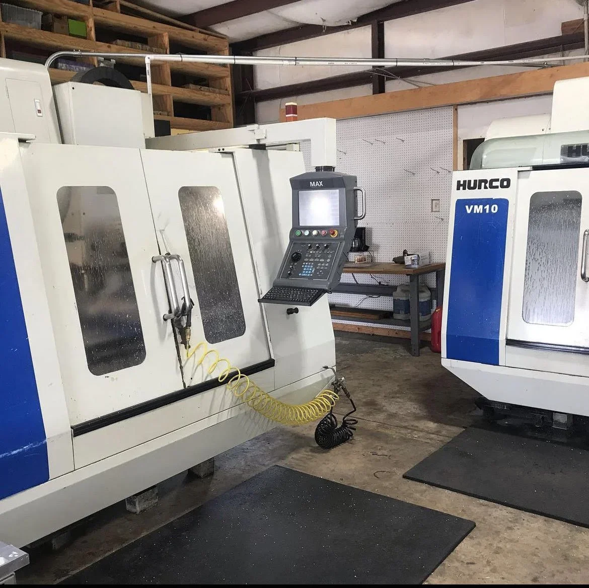 Two CNC machine tools in a workshop, one labeled Hurco VM10, with a computer control panel and safety mats on the floor.