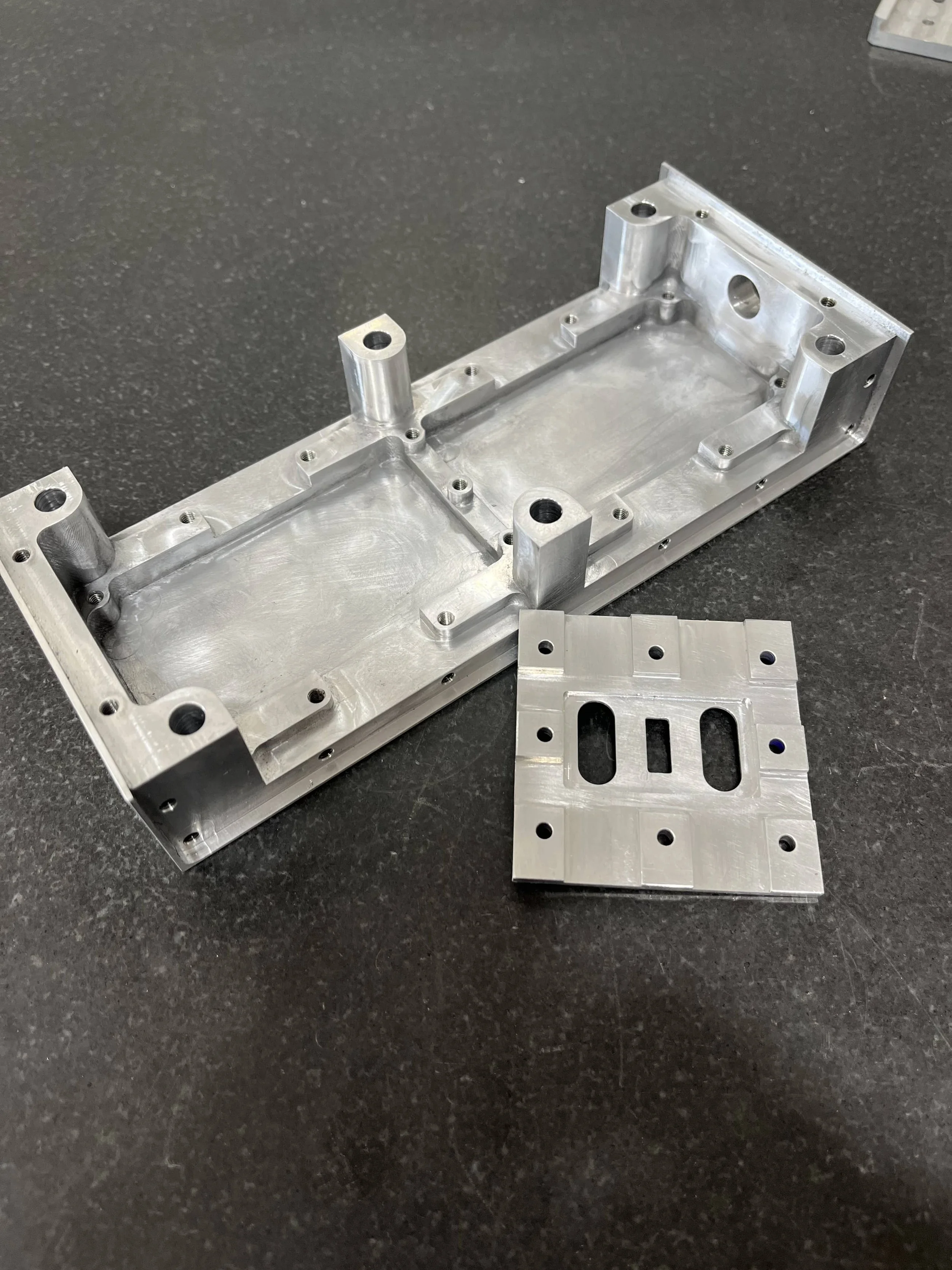 Metal machined components, one rectangular with multiple holes and a larger cutout, and the other smaller with two elongated slots, on a black surface.