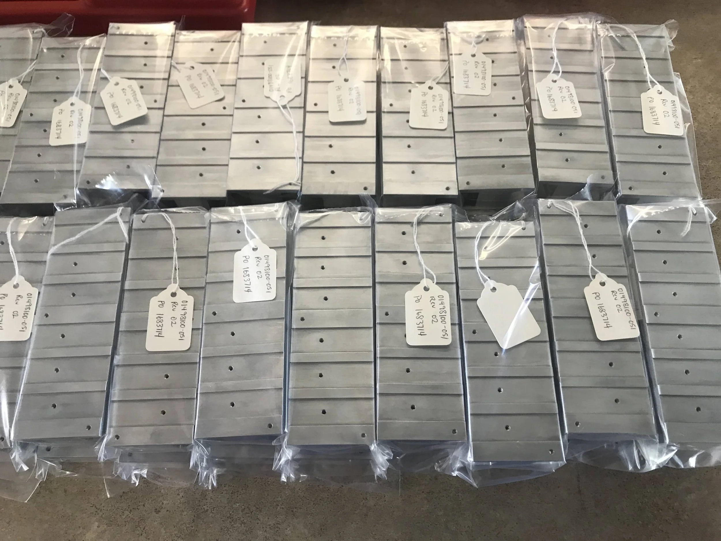 Multiple metal rectangular objects, each wrapped in clear plastic and labeled with white tags, arranged in rows on a flat surface.