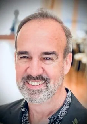 A smiling middle-aged man with a beard and mustache, wearing a dark blazer and patterned shirt, indoors with blurred background.