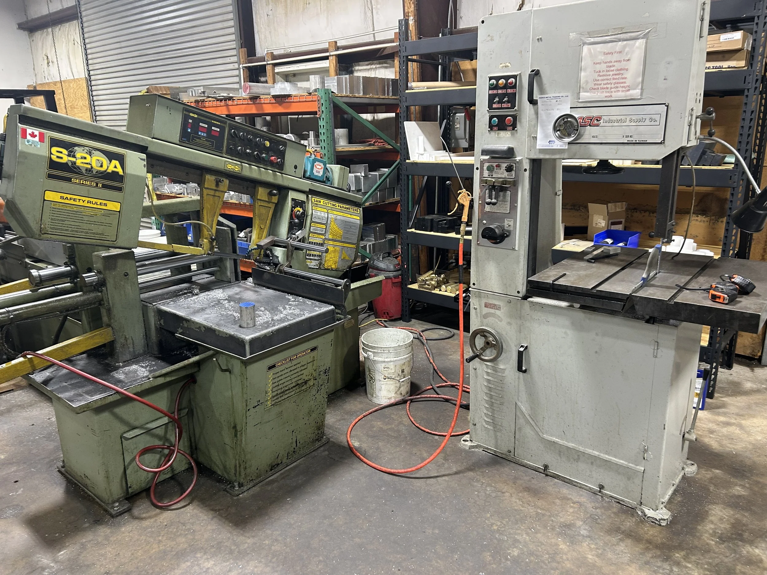 A woodworking workshop with a band saw and a contour surface grinder machine on a concrete floor. Shelving units holding tools and supplies are in the background.