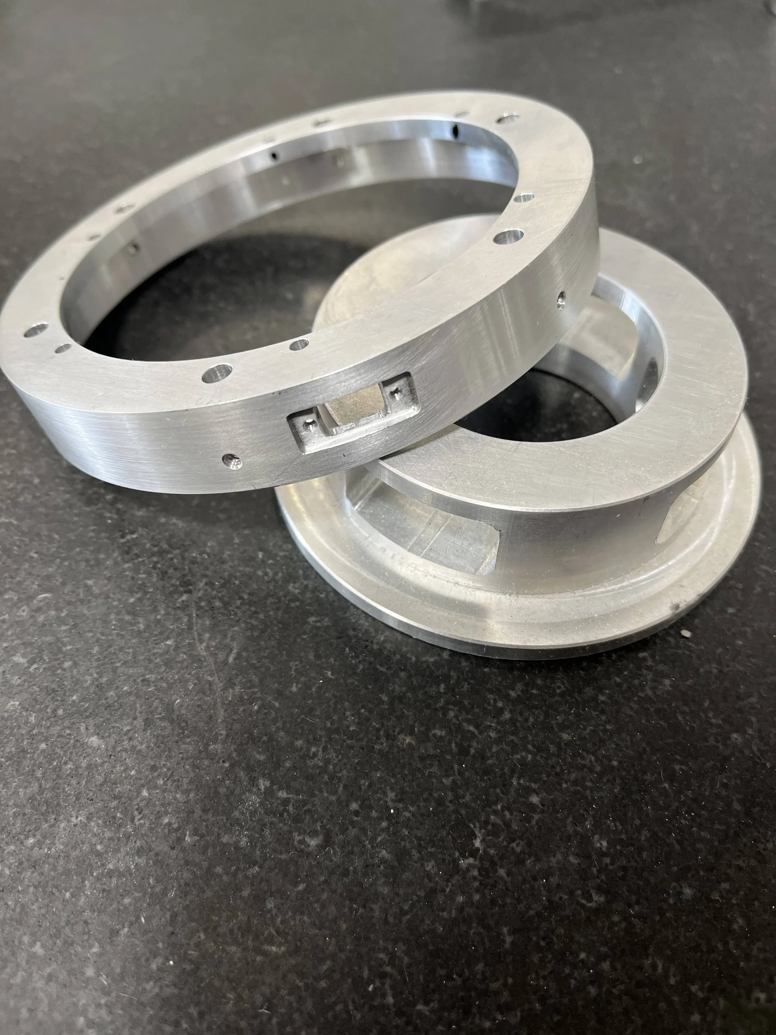 Two machined metal rings with holes and cutouts, placed on a black surface.
