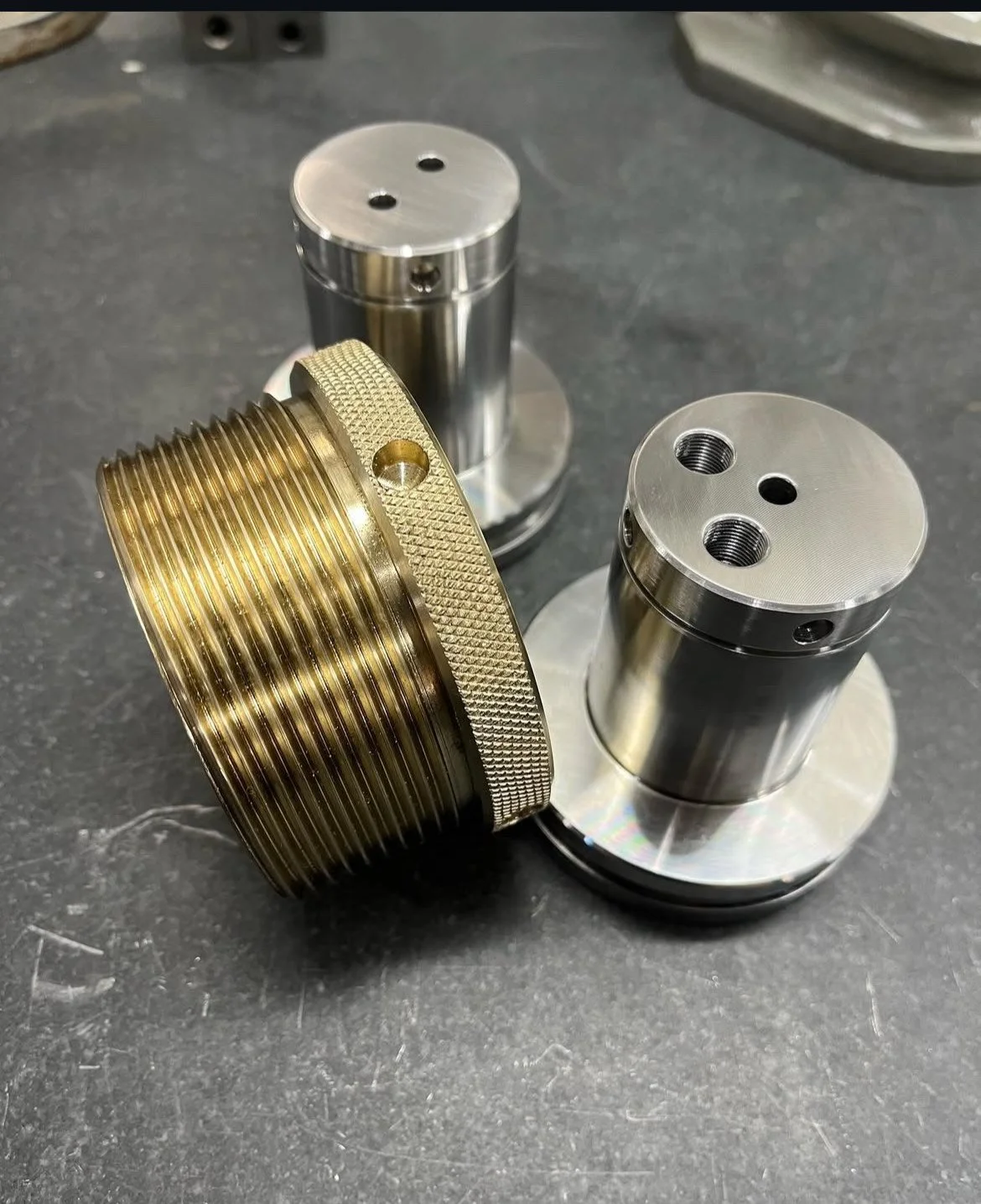 Three machined metal cylindrical components on a dark work surface, two silver and one gold-colored with a knurled edge.