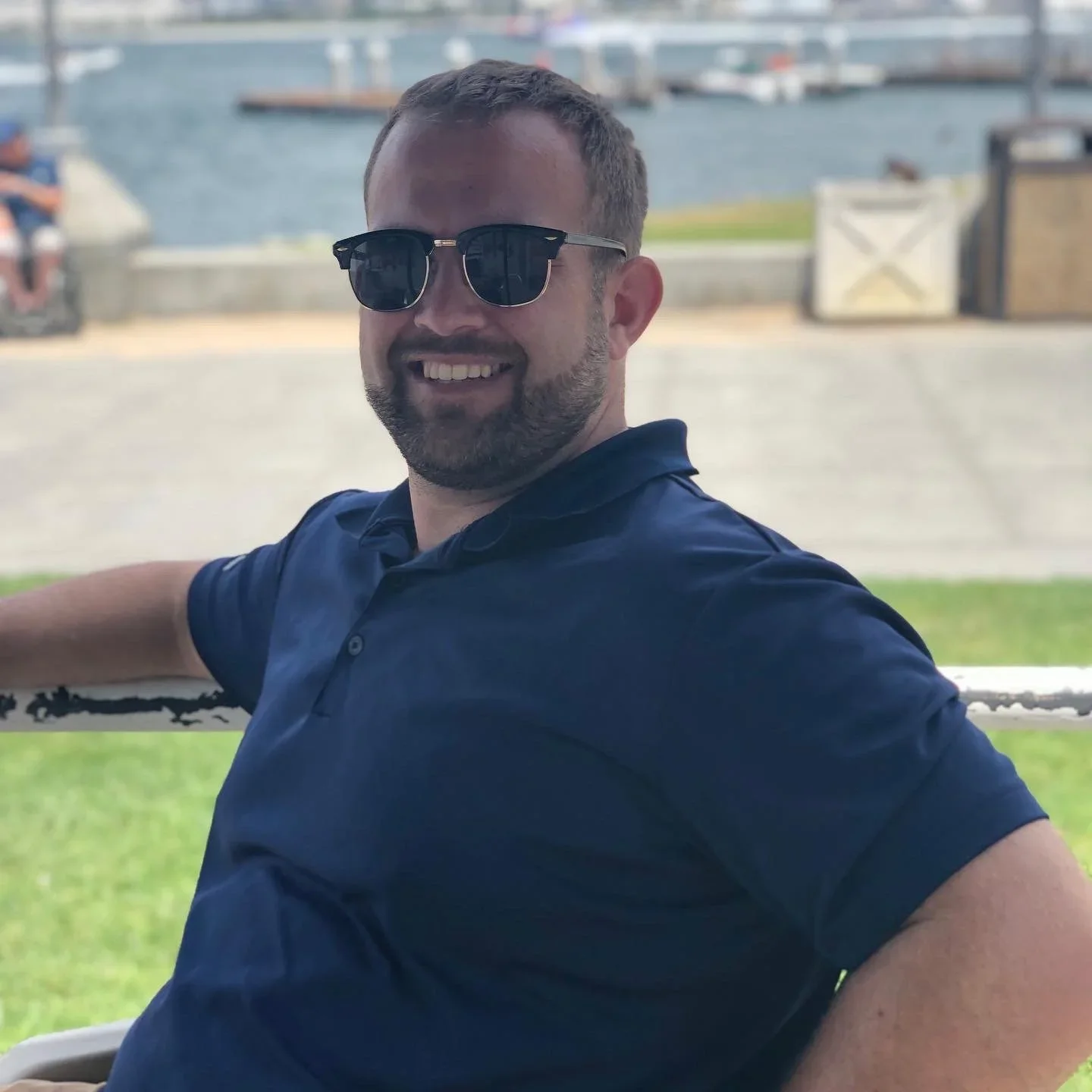 A man with short hair, sunglasses, and a beard, smiling and sitting outdoors near a body of water with a cityscape in the background.