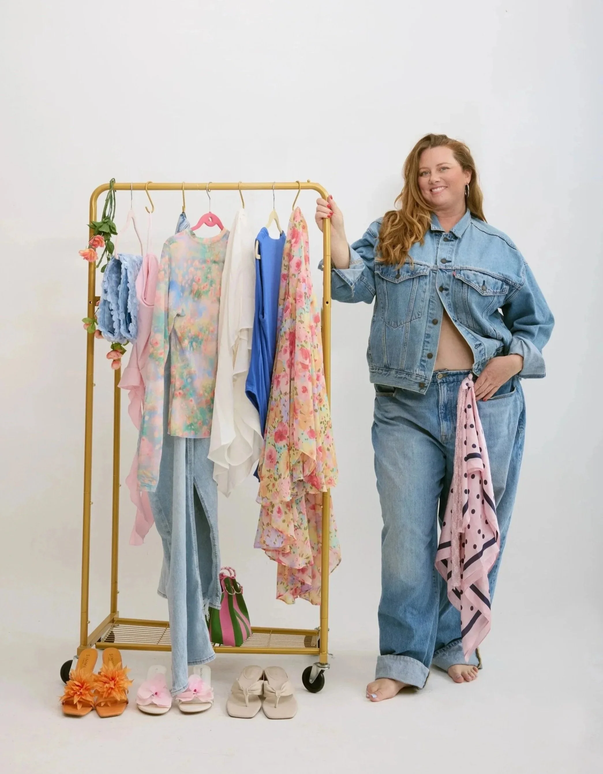 A woman in denim jeans and jacket standing next to a clothing rack with various colorful clothes and shoes, in a white studio.