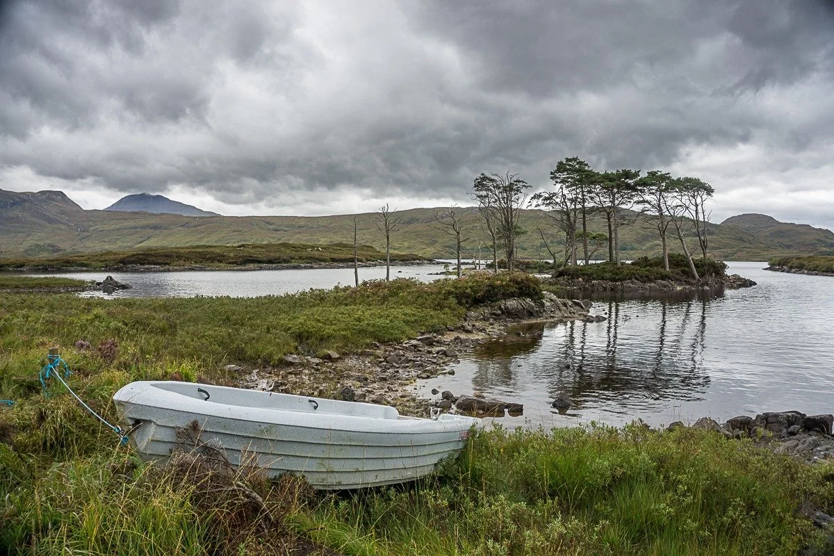 A small white boat resting on grassy land near a lake with a small wooded island and hills in the background, under a cloudy sky.
