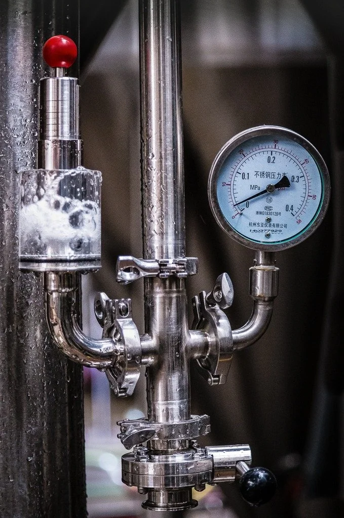 Close-up of stainless steel brewing or distilling equipment with a pressure gauge reading approximately 0.2 MPa, and water droplets on the surface.