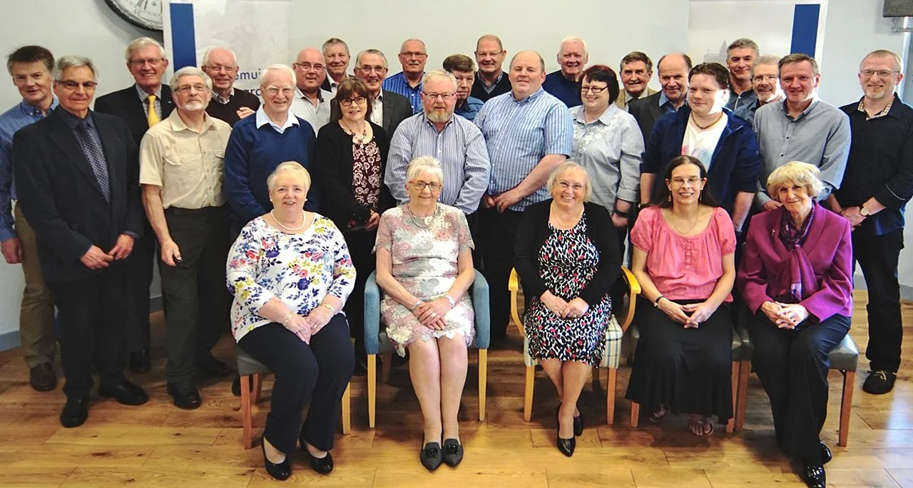 Group photo of approximately 25 middle-aged to elderly adults, standing and seated indoors, posed for a formal or semi-formal celebration or gathering.