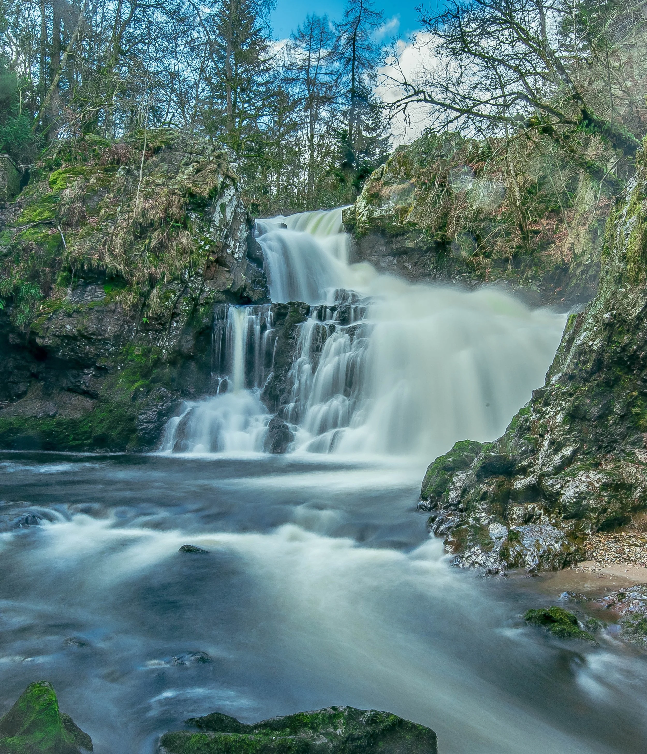 A cascading waterfall flowing over rocks in a forest setting with trees and a partly cloudy sky in the background.