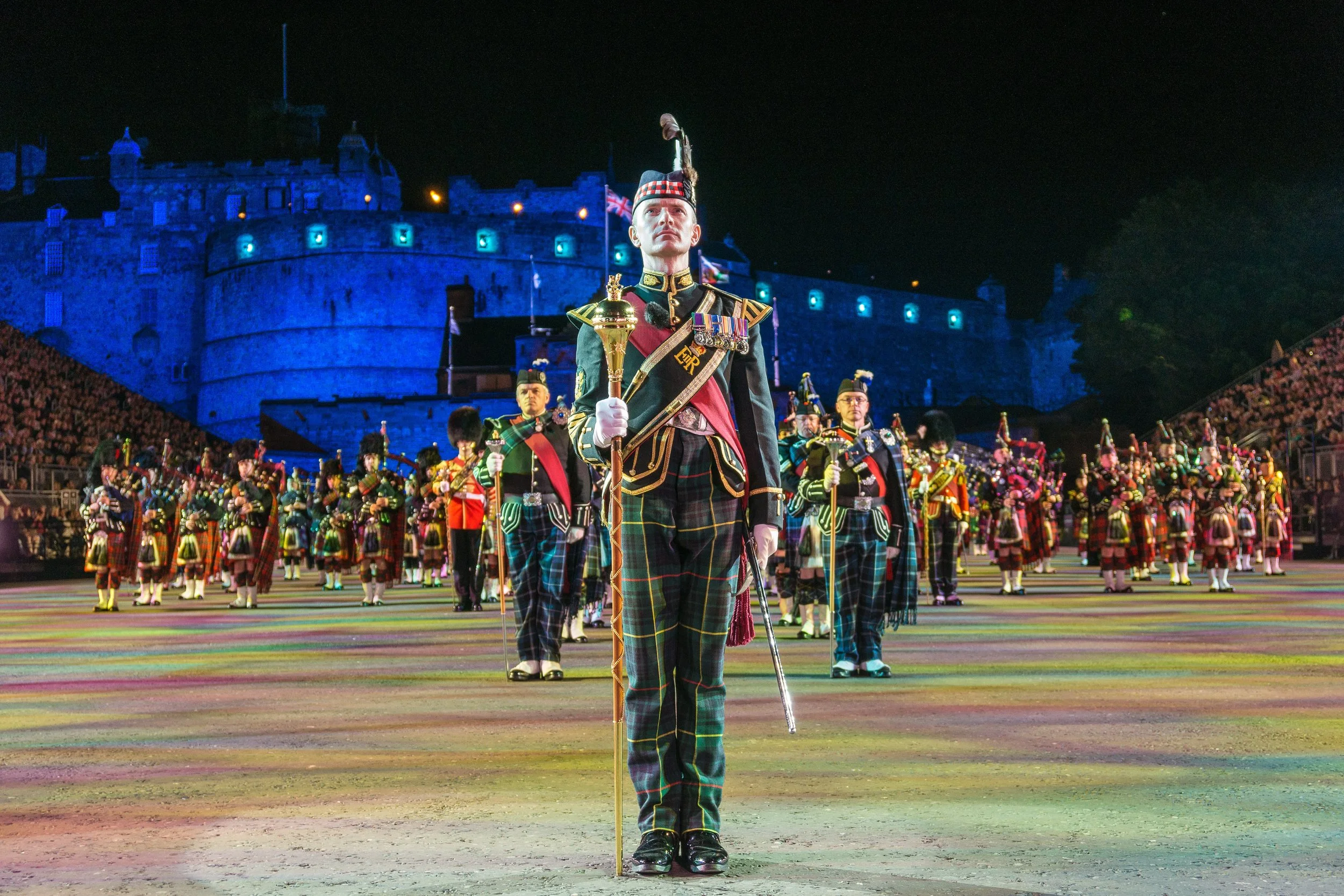 A group of soldiers in dress uniform, including a soldier in the foreground holding a ceremonial baton, standing at attention in front of a castle illuminated with blue lights at night.