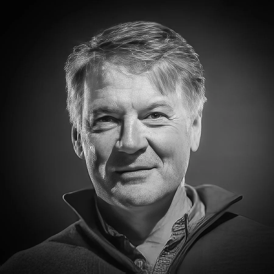 Black and white portrait of a middle-aged man with short, light-colored hair, wearing a collared shirt and zip-up sweater.