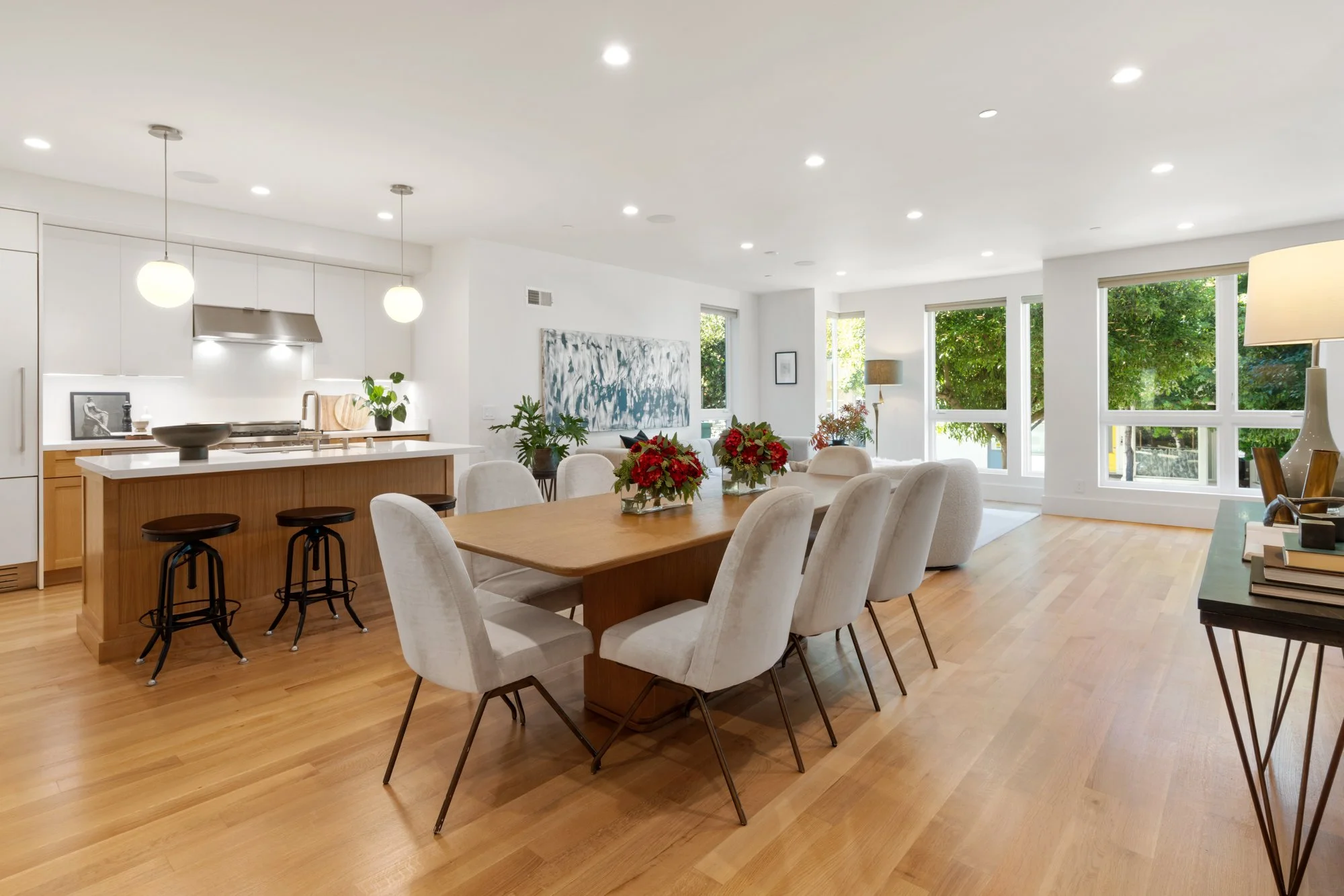 Open-concept living and dining area with hardwood floors, white walls, large windows, a dining table with eight chairs, and a kitchen island with three barstools.