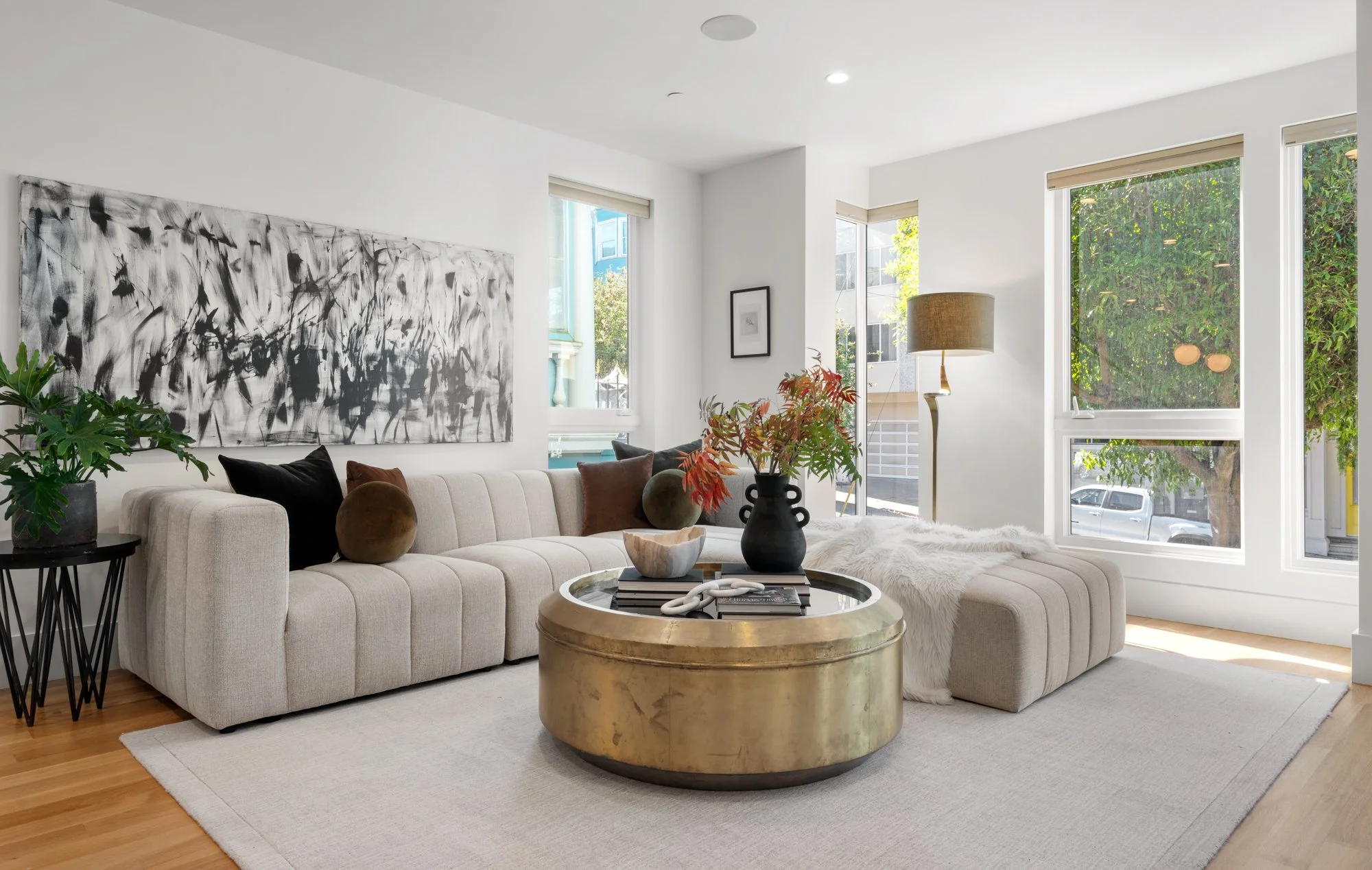 Bright living room with white walls, large windows, beige sectional sofa with pillows, black side table with potted plant, gold round coffee table with vase of flowers, abstract black and white art on wall, floor lamp, and light hardwood flooring.