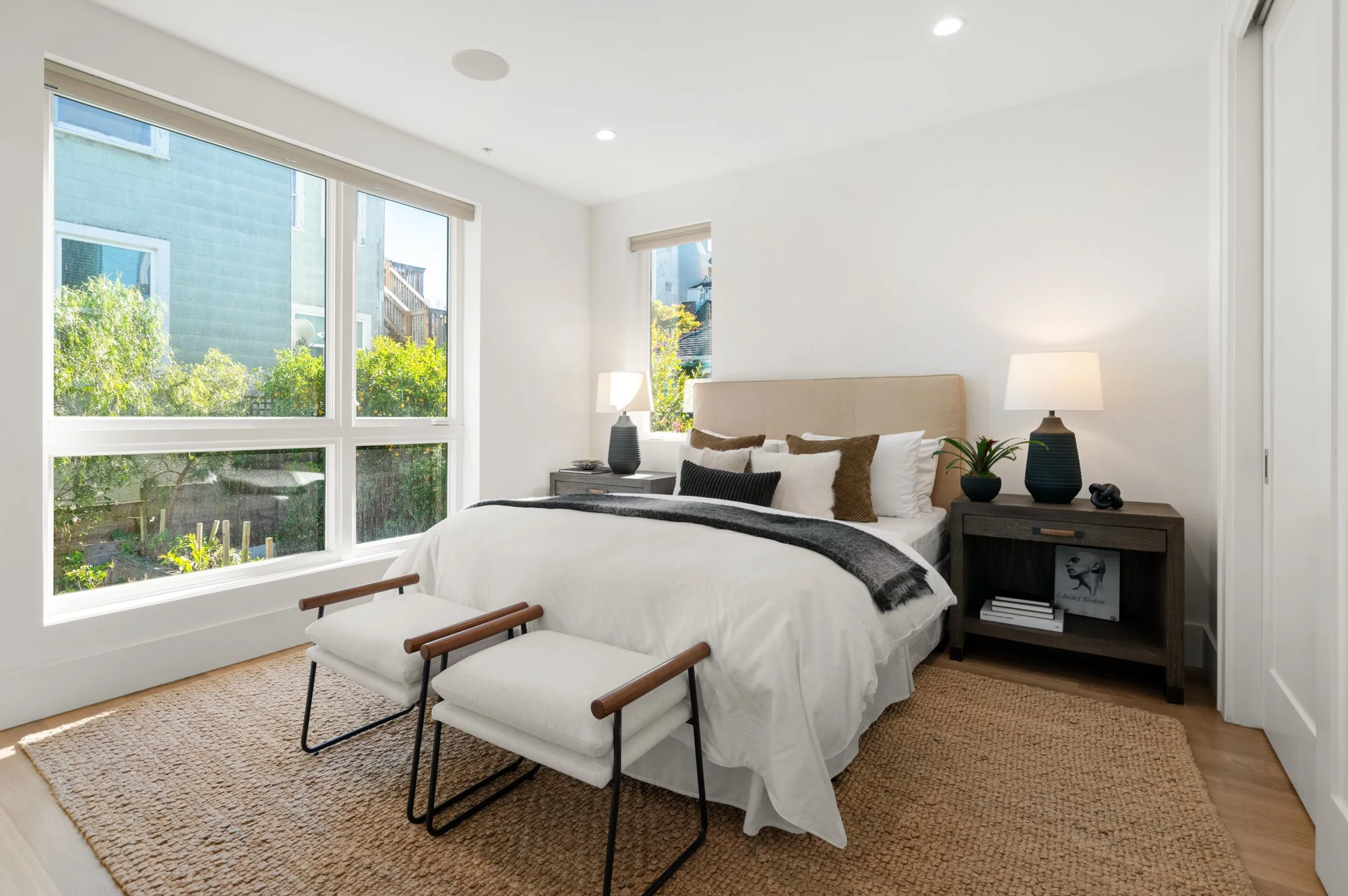 A bright bedroom featuring a bed with white bedding, brown and black pillows, and a beige headboard. There are two dark wooden nightstands with lamps, a plant, and decorative items. Large windows let in natural light, revealing a garden outside. A wo