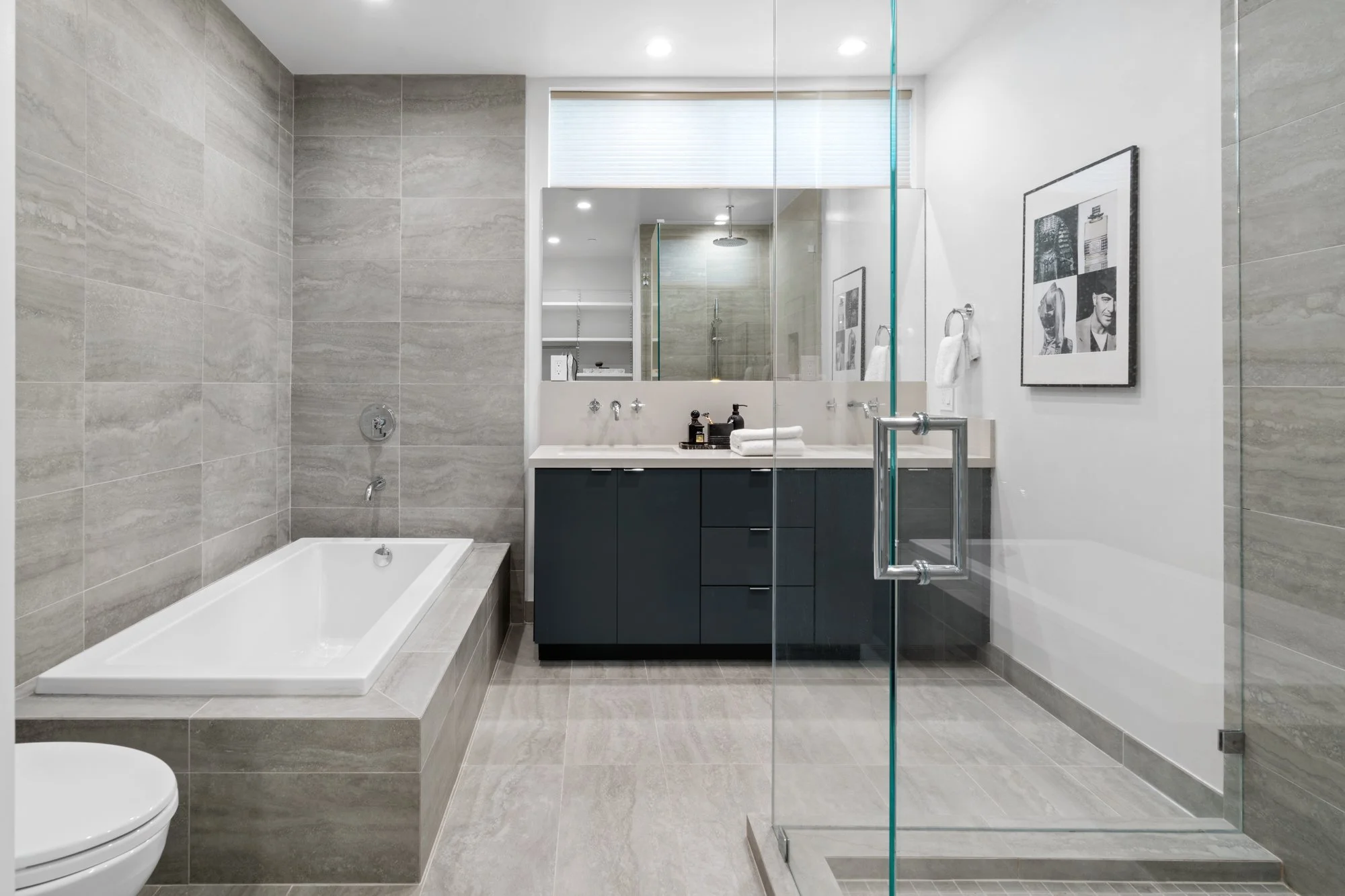 Modern bathroom with gray tiled walls and floor, a bathtub on the left, a glass shower enclosure on the right, and a dark blue vanity with a white countertop and large mirror above. The bathroom has white walls, wall-mounted towels, and a framed blac