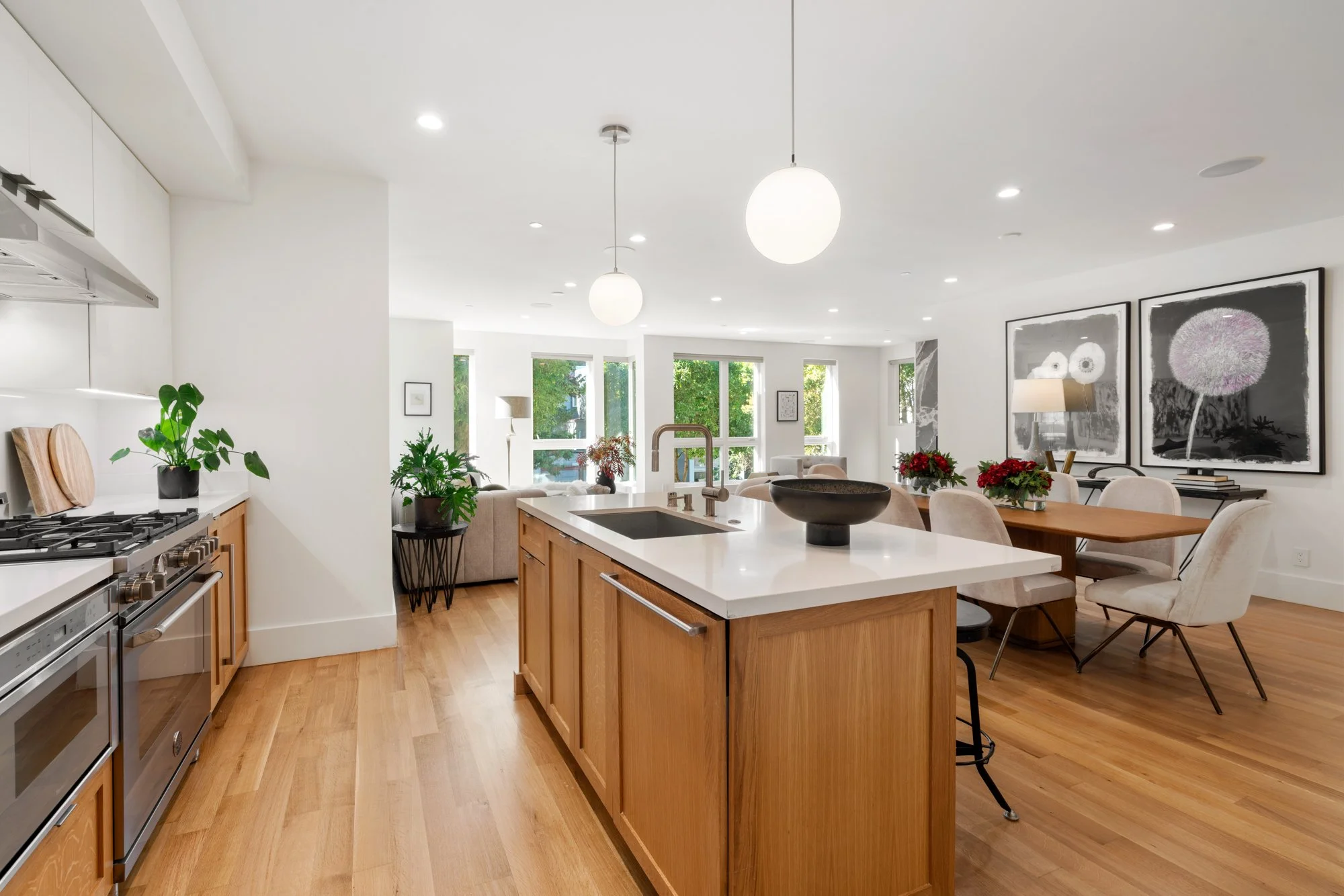 Open-concept kitchen and dining area with white walls, wooden flooring, a kitchen island with a white countertop, plants, and modern wall art.