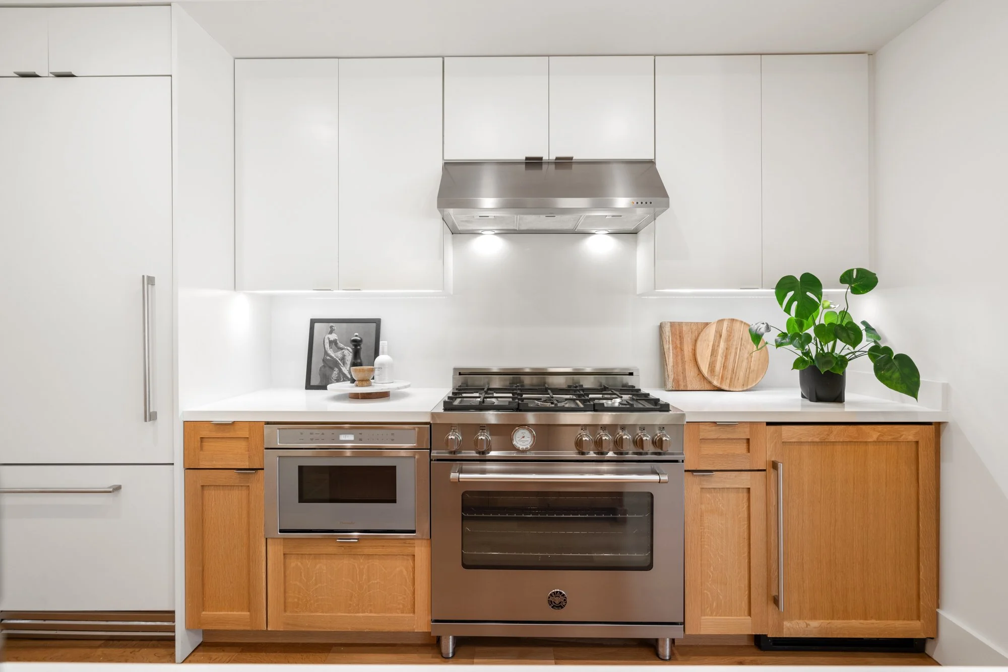 Modern kitchen with white cabinets, a stainless steel gas oven and range, a microwave, a refrigerator, wooden cabinetry, a plant, and decorative items on the countertop.
