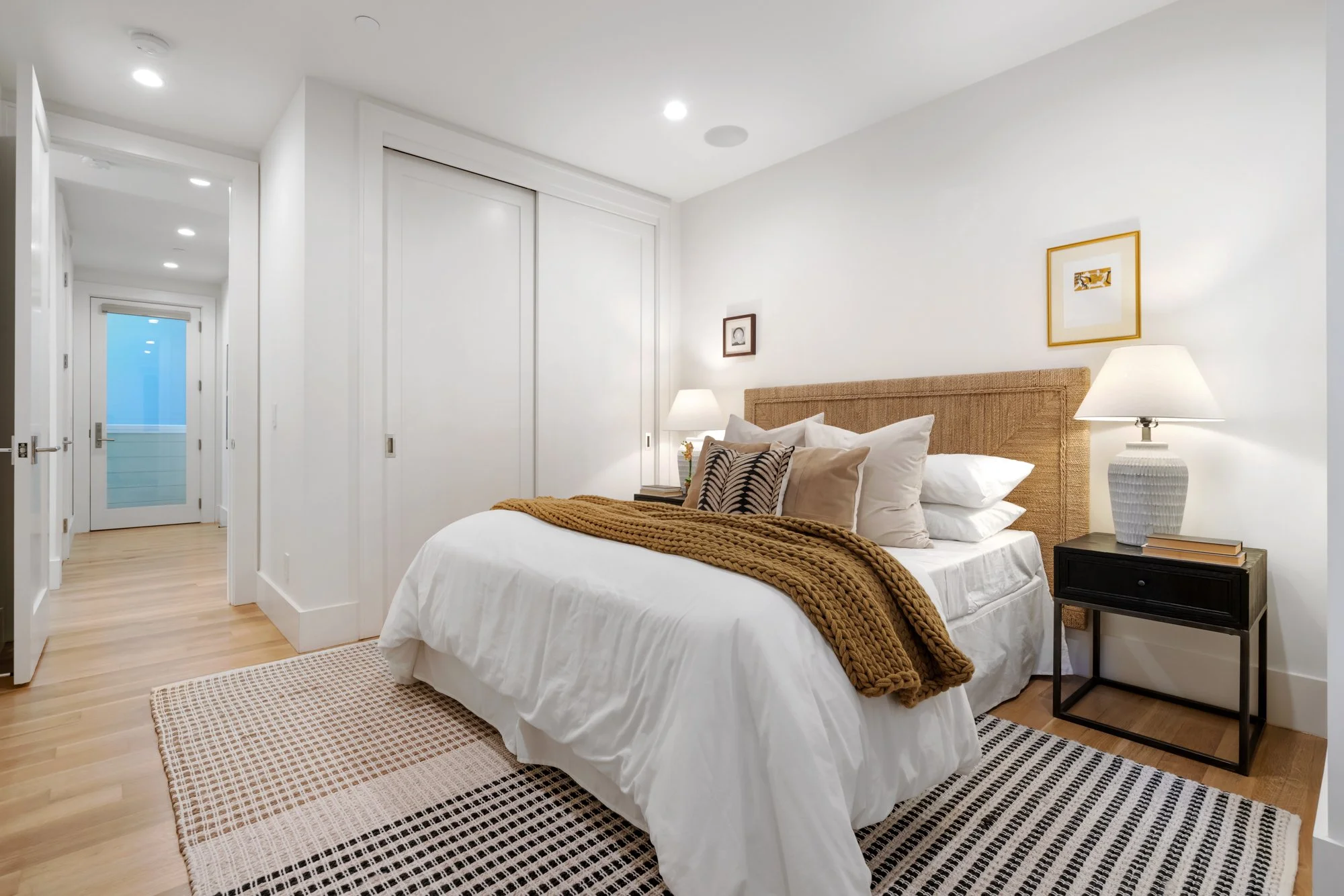 A bedroom with a bed, pillows, and a brown throw blanket; nightstand with a lamp and books; hardwood floor and patterned area rug; white walls and ceiling, with a door and closet.