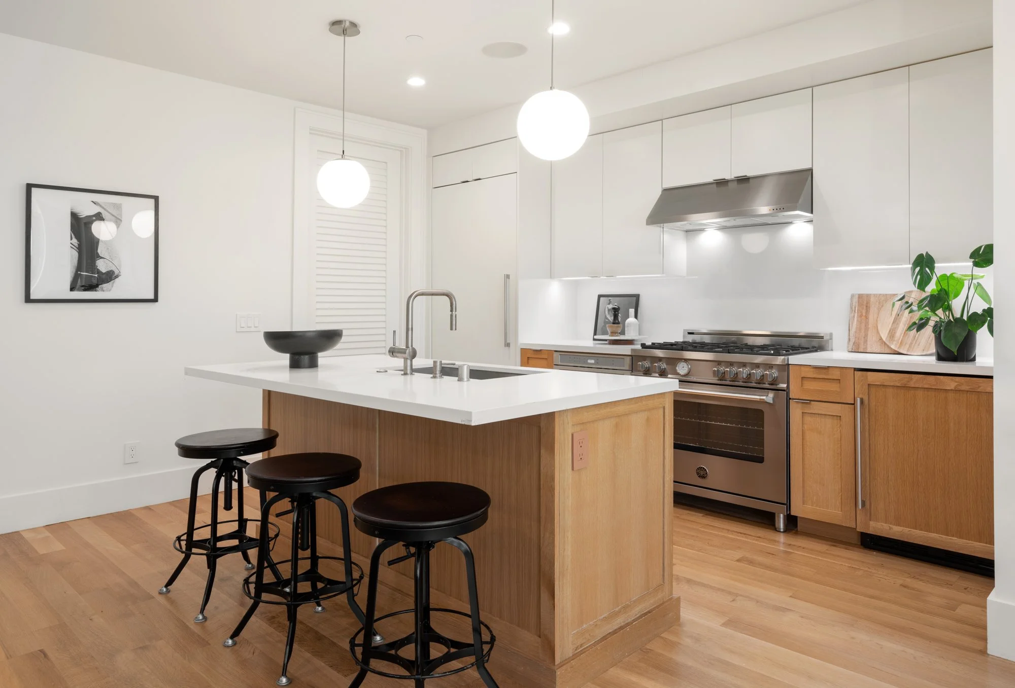 Modern kitchen with white walls, wooden cabinets, and a central island with a white countertop. There are three black barstools at the island. The kitchen has stainless steel appliances, including a range and hood. Hanging above the island are two ro