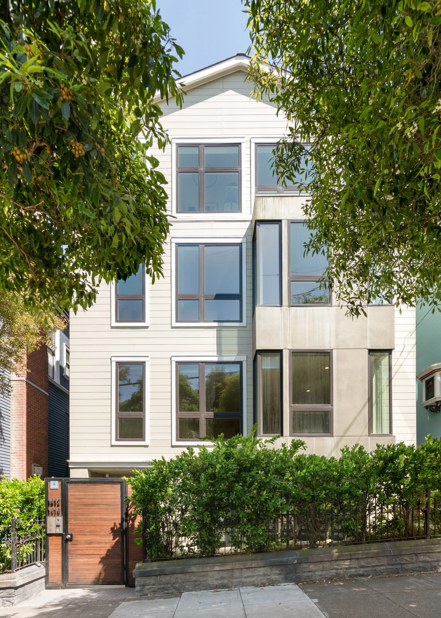 Modern multi-story residential building with large windows, surrounded by green trees and shrubs, with a metal and wood gate at the front.