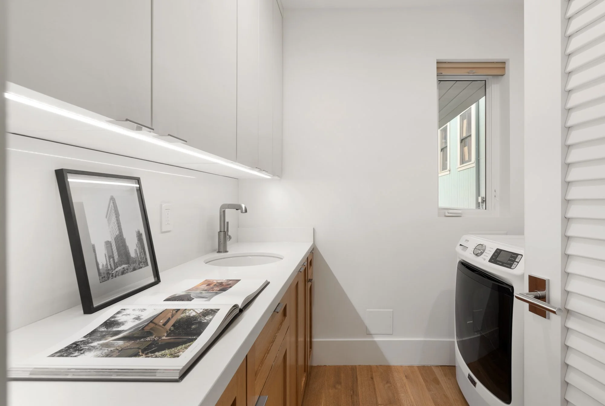 A laundry room with white walls, wooden cabinets, a white countertop, a framed black-and-white cityscape photo, an open photo book, a small round sink, a silver faucet, a small window with a blind, and a white washing machine.