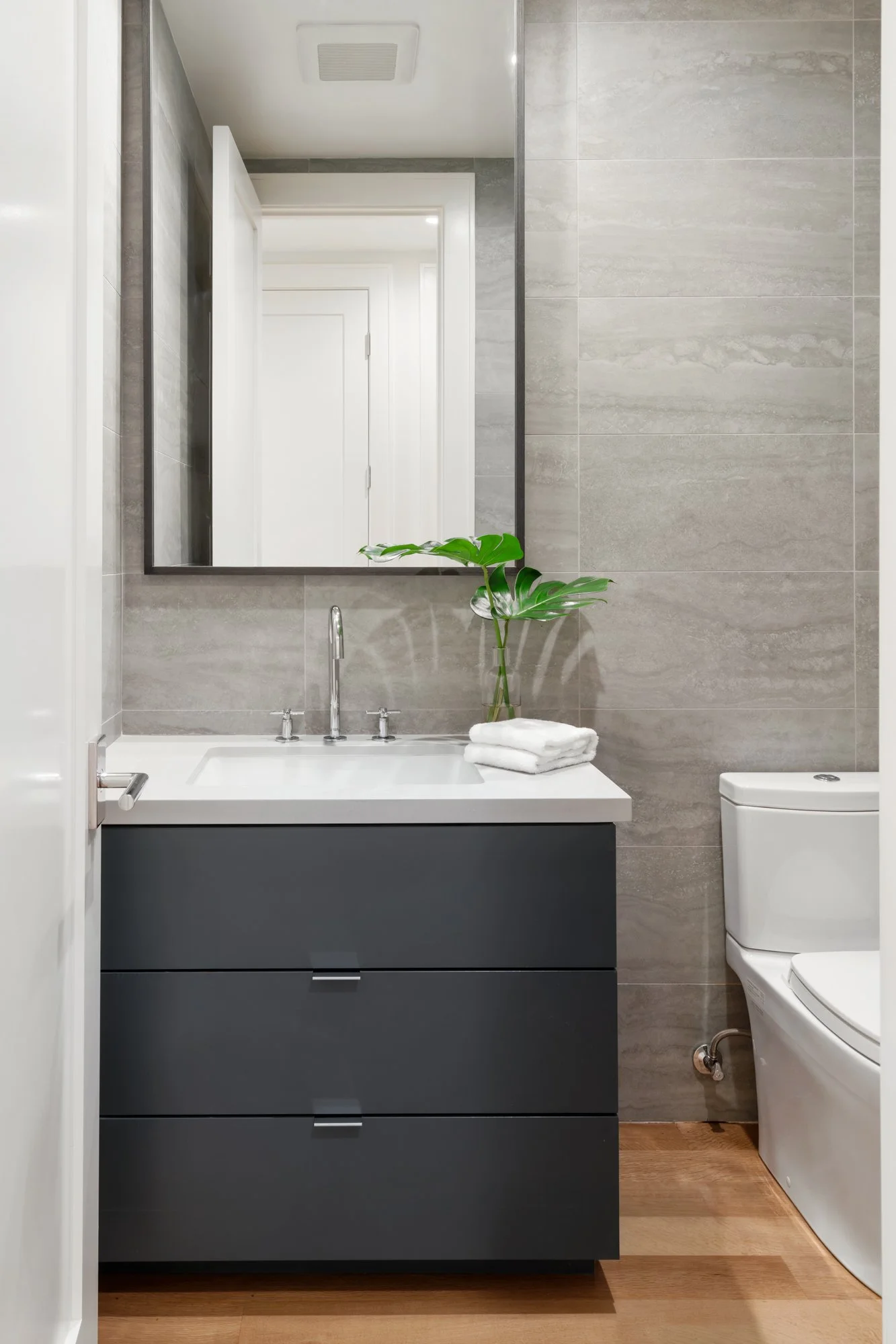 Modern bathroom with a wall-mounted mirror, a gray vanity with white countertop, a faucet, a green plant, rolled white towels, and a toilet against a tiled wall.