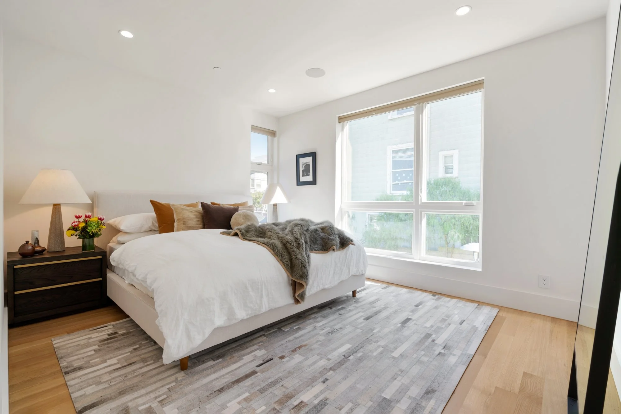 A bedroom with a white bed, multiple pillows, a bedside table with a lamp and flowers, large windows allowing natural light, and a patterned area rug on a wooden floor.