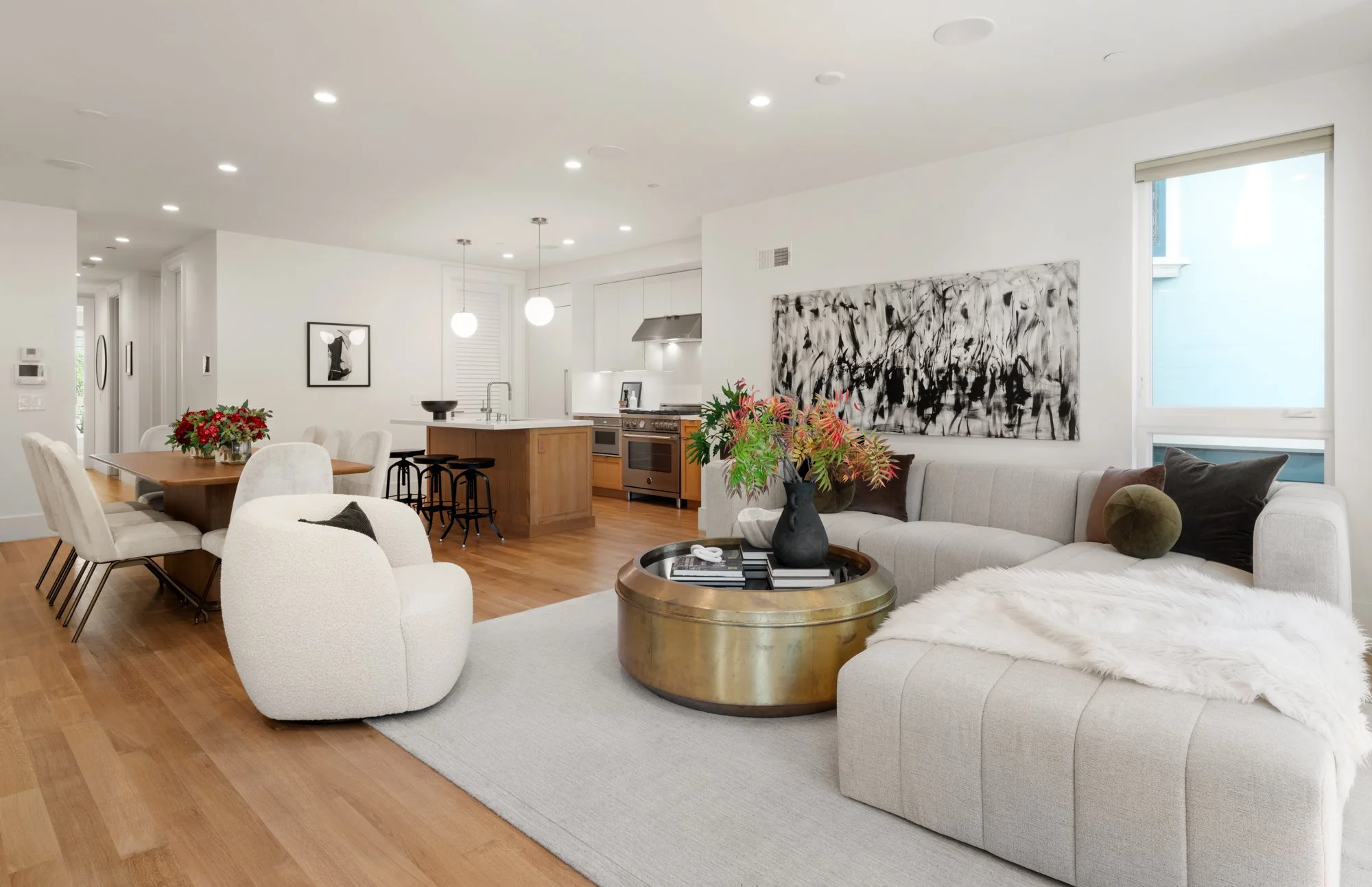 Modern open-concept living room and kitchen with white walls, wood flooring, beige sofa, white armchair, gold coffee table, black and green decorative pillows, abstract black and white wall art, large window, and dining table with chairs.