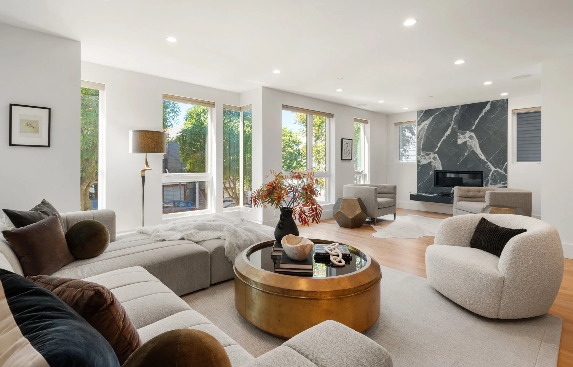 Bright living room with large windows, modern furniture, a gold coffee table, a black vase with red foliage, a fireplace with a black and white marble surround, and hardwood floors.