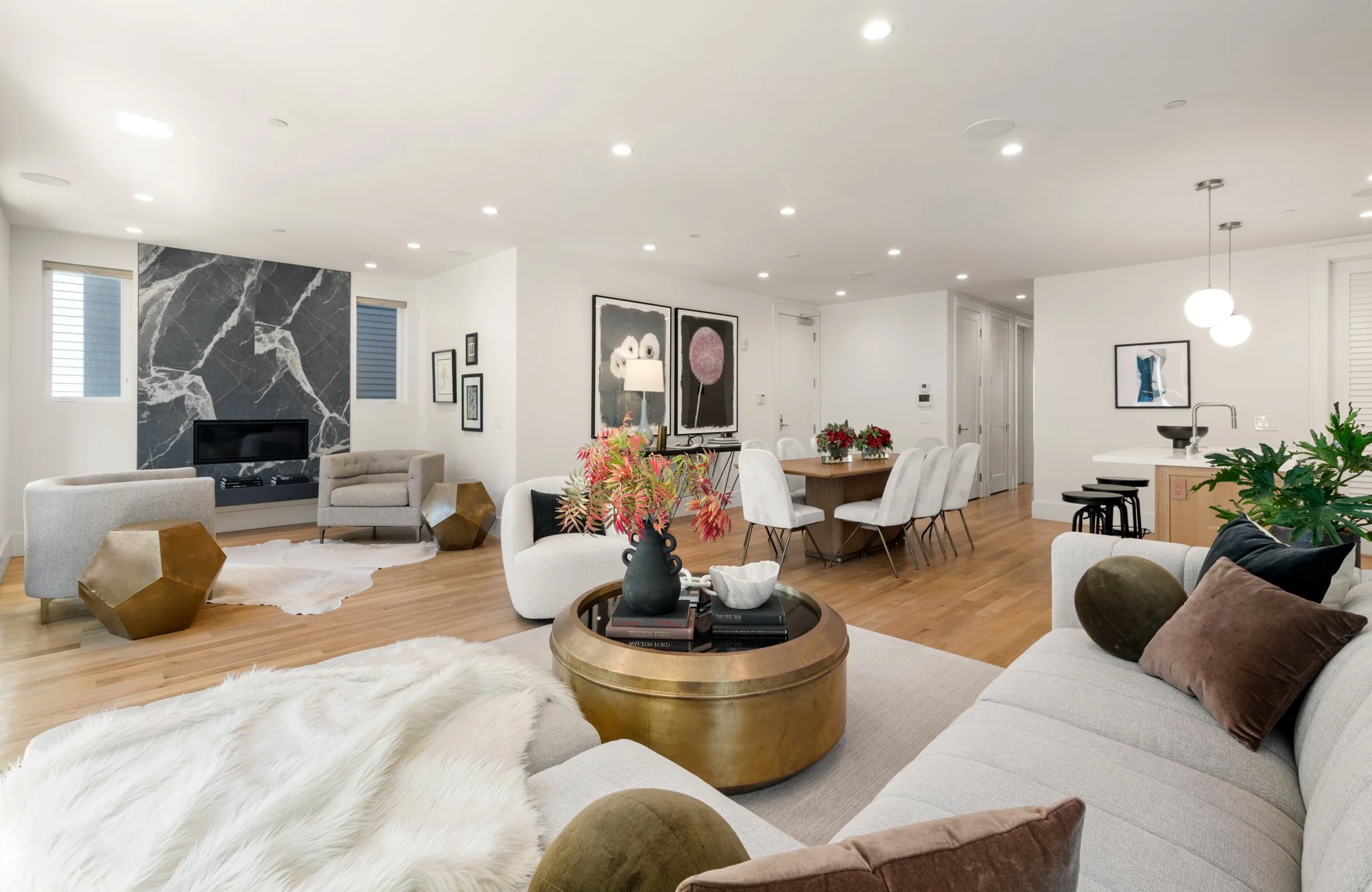 Modern living room with white walls, wooden flooring, and ample seating including a sofa and armchairs. Features a fireplace with black marble surround, decorative objects, and artwork on the walls. There is a dining table with white chairs, a small 