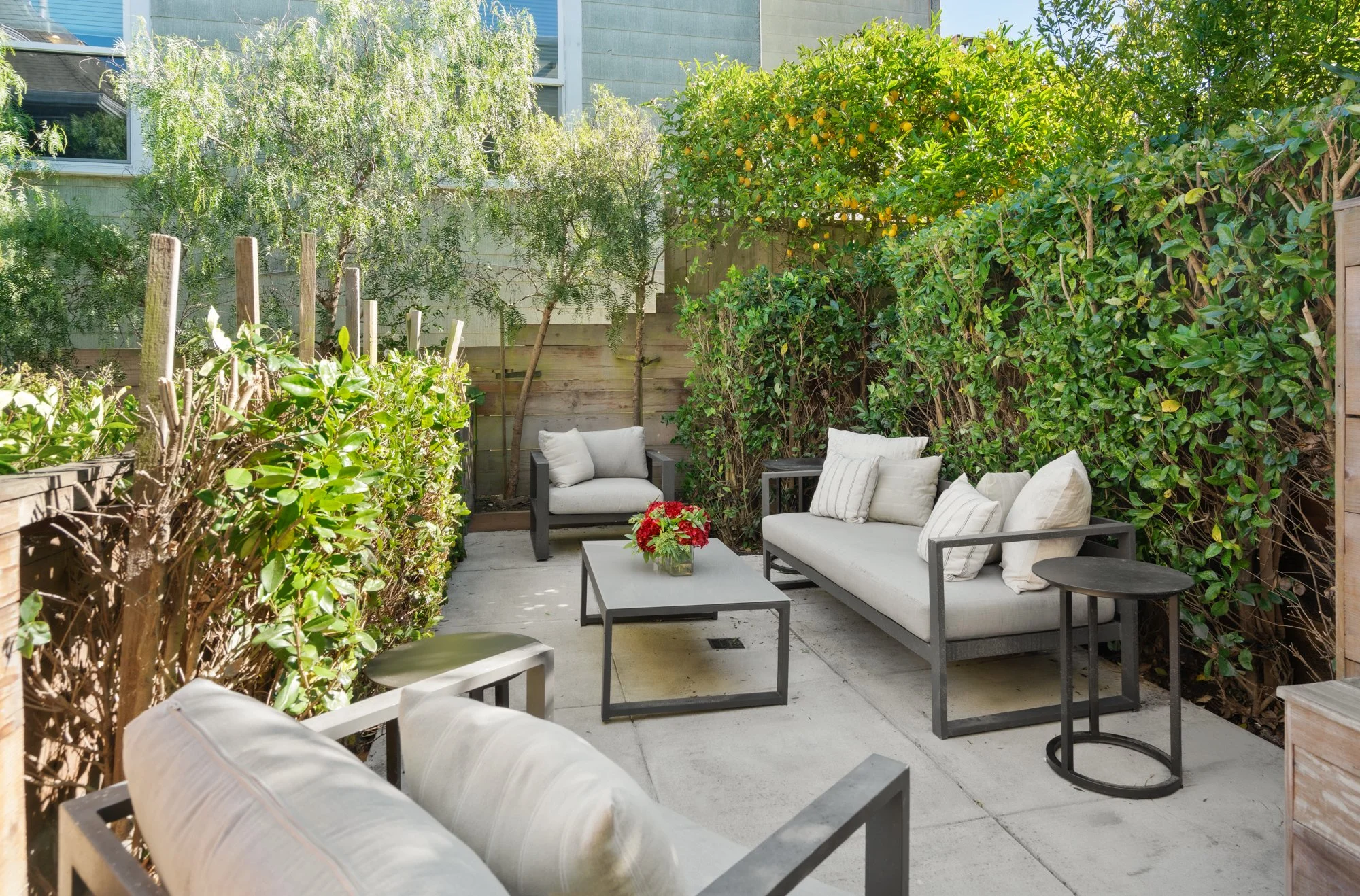 Outdoor patio with gray and black furniture, seating cushions, a coffee table with a red flower arrangement, and lush green bushes and trees surrounding the space.
