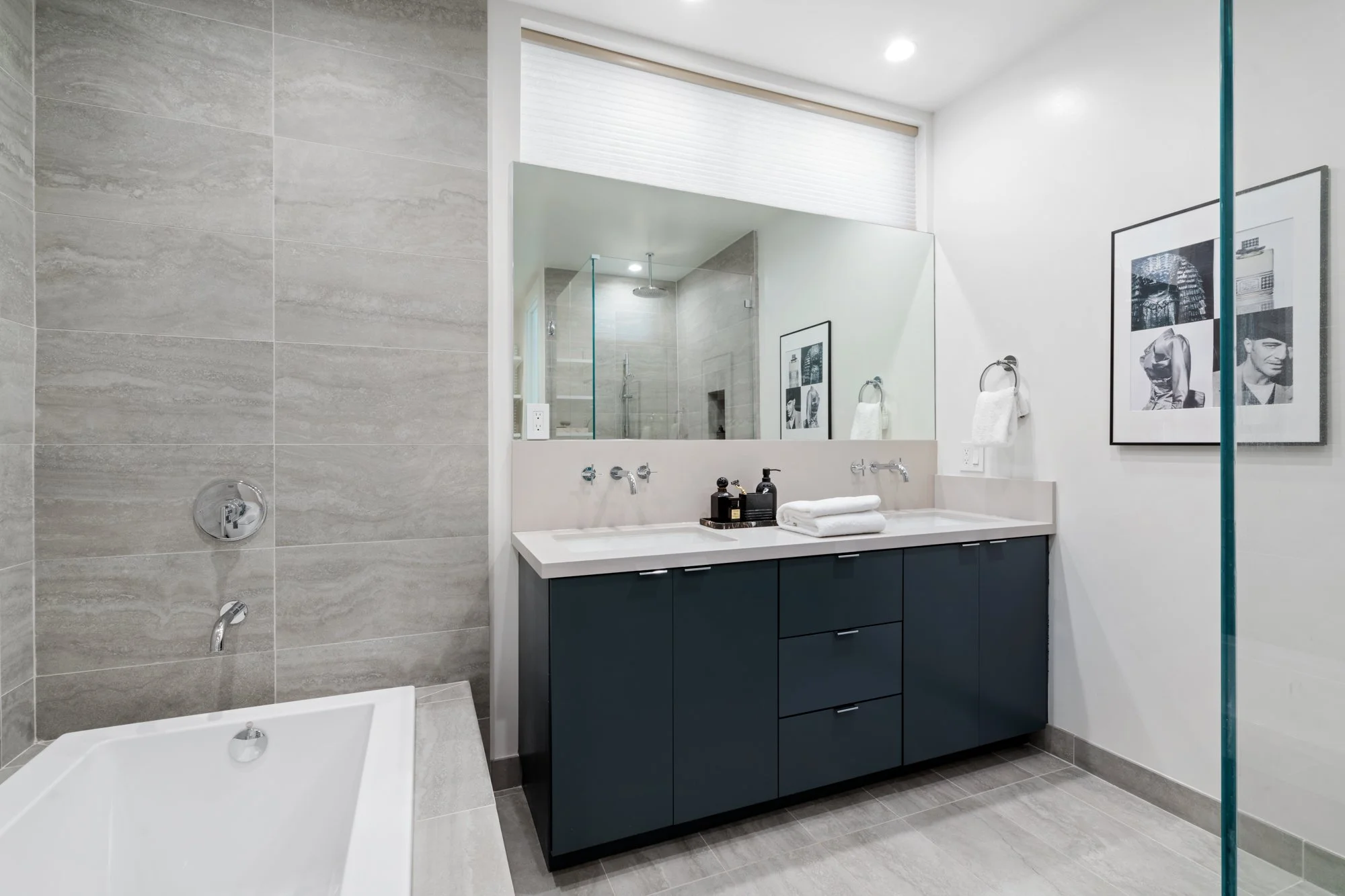 Modern bathroom with a large mirror, dual sinks, dark blue vanity, bathtub, framed black and white photographs, and a glass shower enclosure.