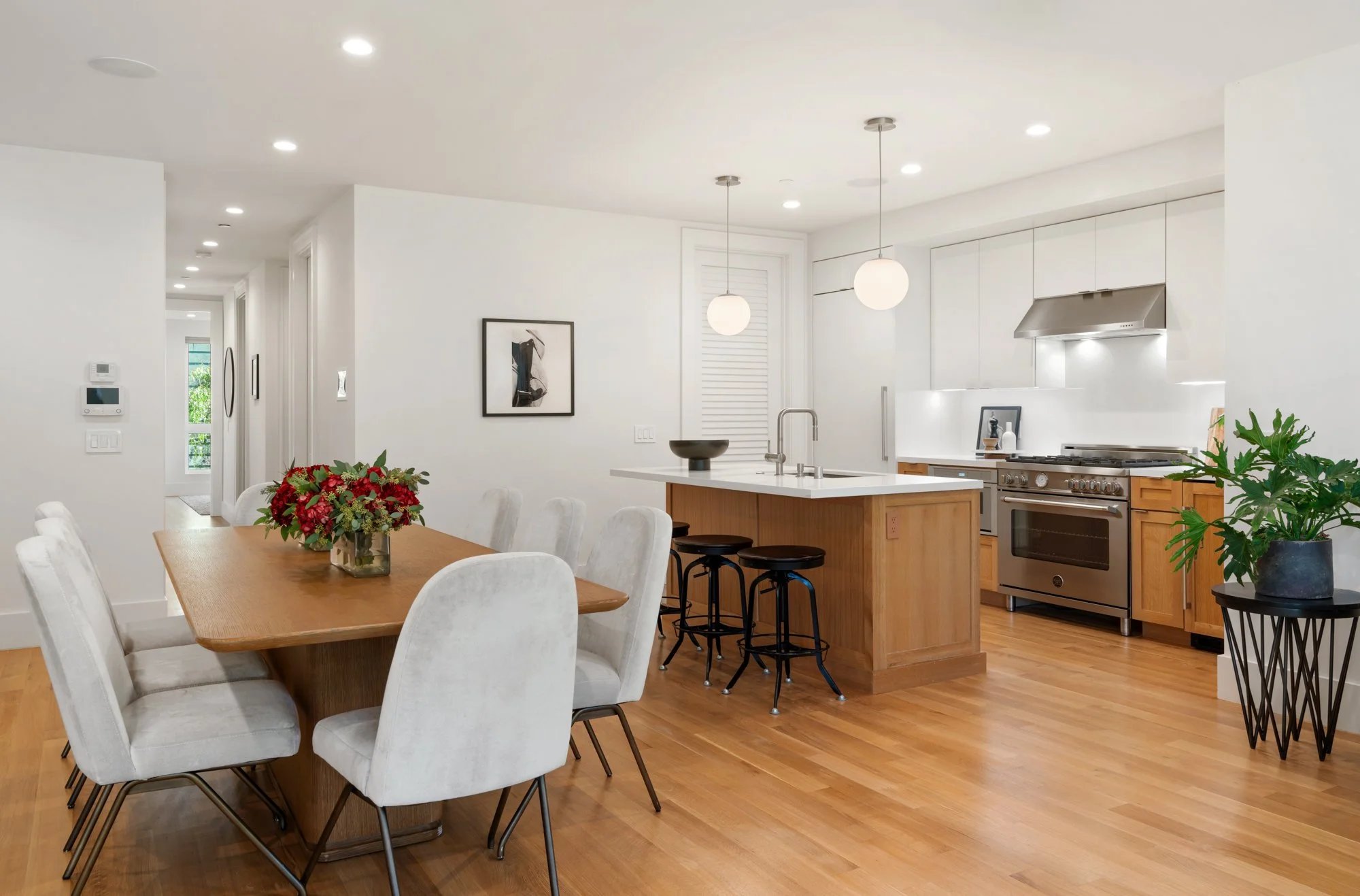 Modern kitchen and dining area with a wooden dining table surrounded by white upholstered chairs, a kitchen island with stools, and stainless steel appliances, including a stove and range hood. Decor includes a framed art piece on the wall, a vase wi