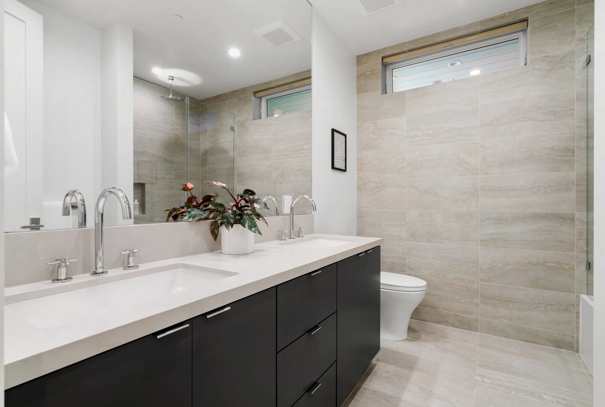 Modern bathroom with beige tiled walls and floors, a large mirror, a white countertop with two sinks, black cabinetry, a potted plant, a toilet, and small windows near the ceiling.