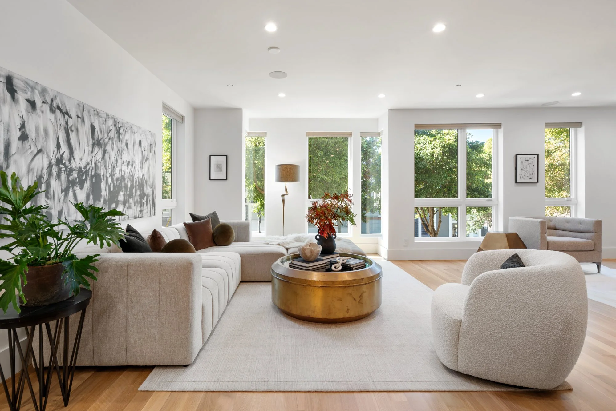 Modern living room with white walls, large windows, a beige sectional sofa with brown and black cushions, a white armchair, a round gold coffee table, a potted plant, a side table, and framed artwork, with wooden flooring and natural light.