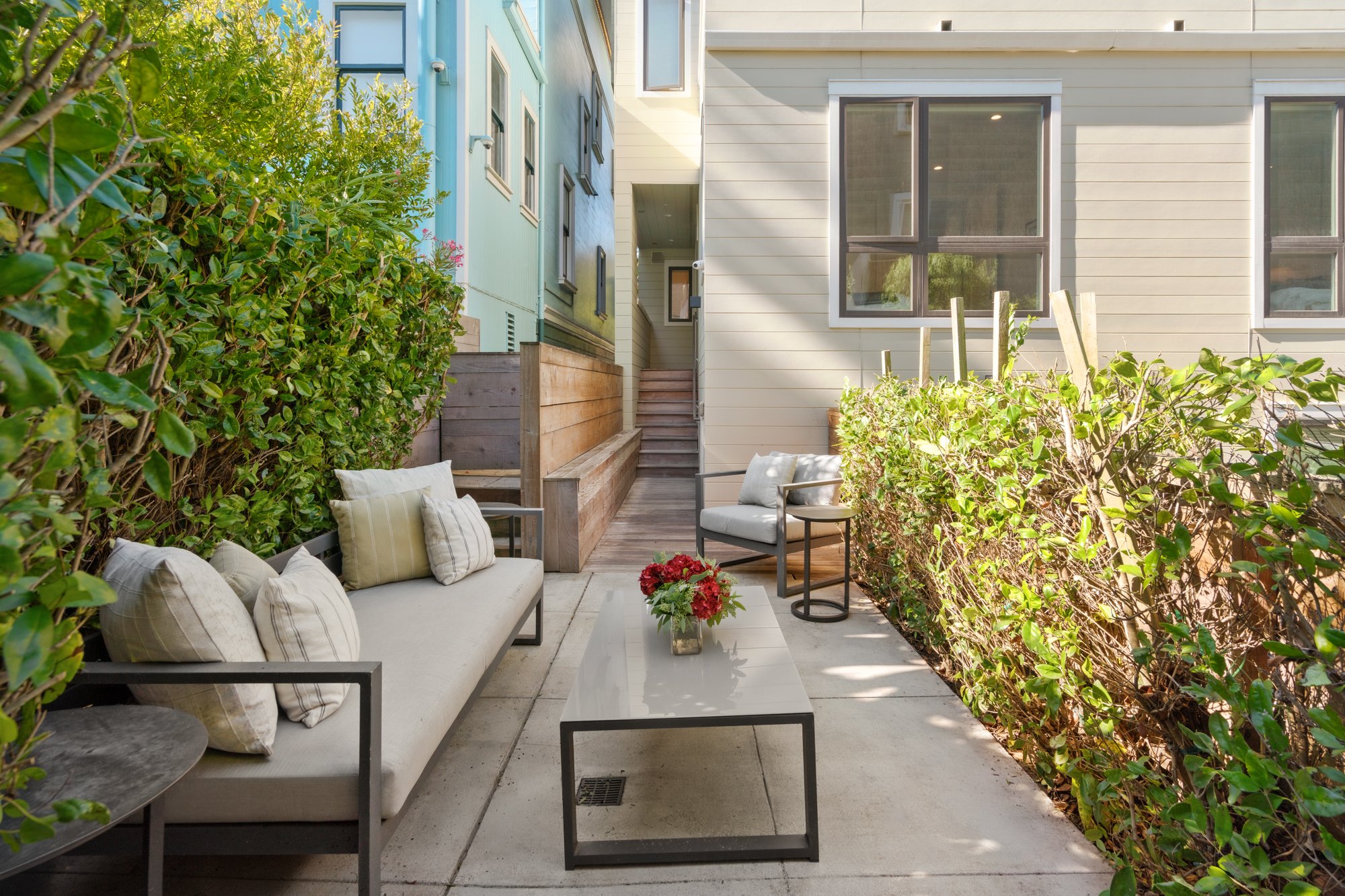 Outdoor patio area with a sofa and an armchair surrounded by green bushes, a coffee table with a vase of red flowers, and neighboring houses in the background.