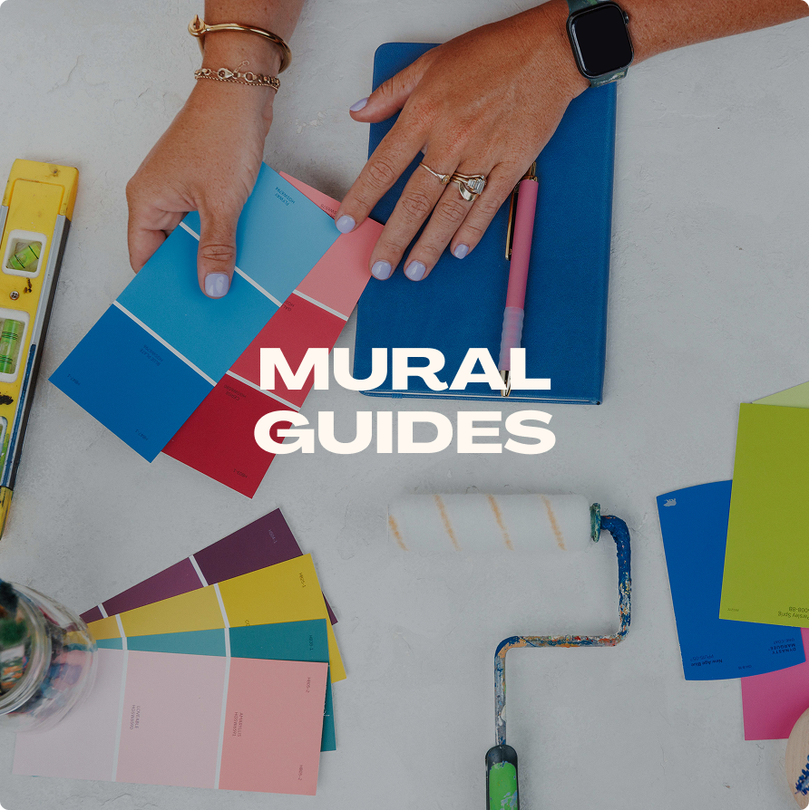 Hands working with color swatches, notepad, paint roller, and stationery on a white table, with the text 'MURAL GUIDES' overlaid.