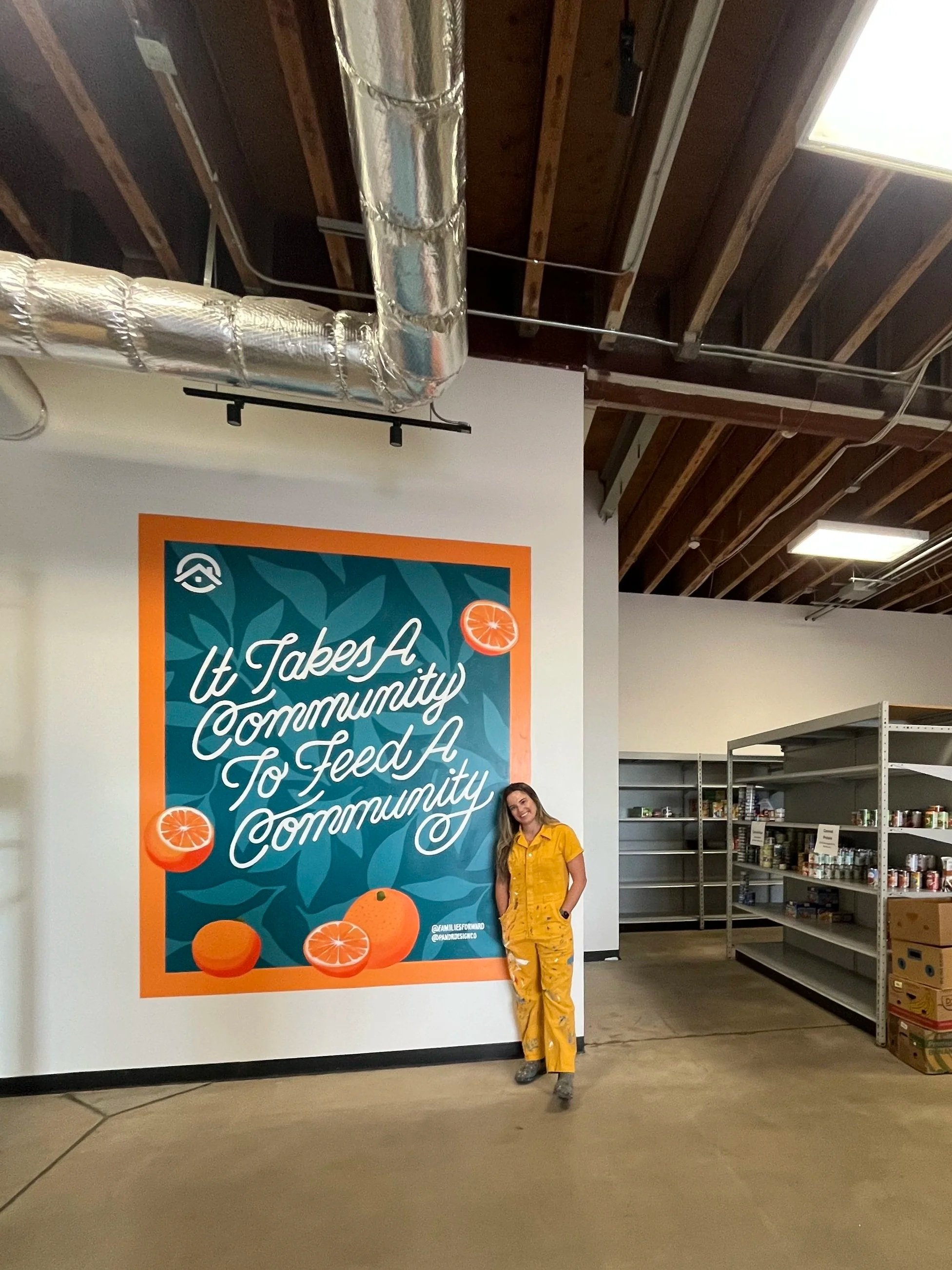 A photo of a painter standing in front of her mural of the words "it takes a community to feed a community" over a blue floral background, with an orange border and orange slices painted around the edges.