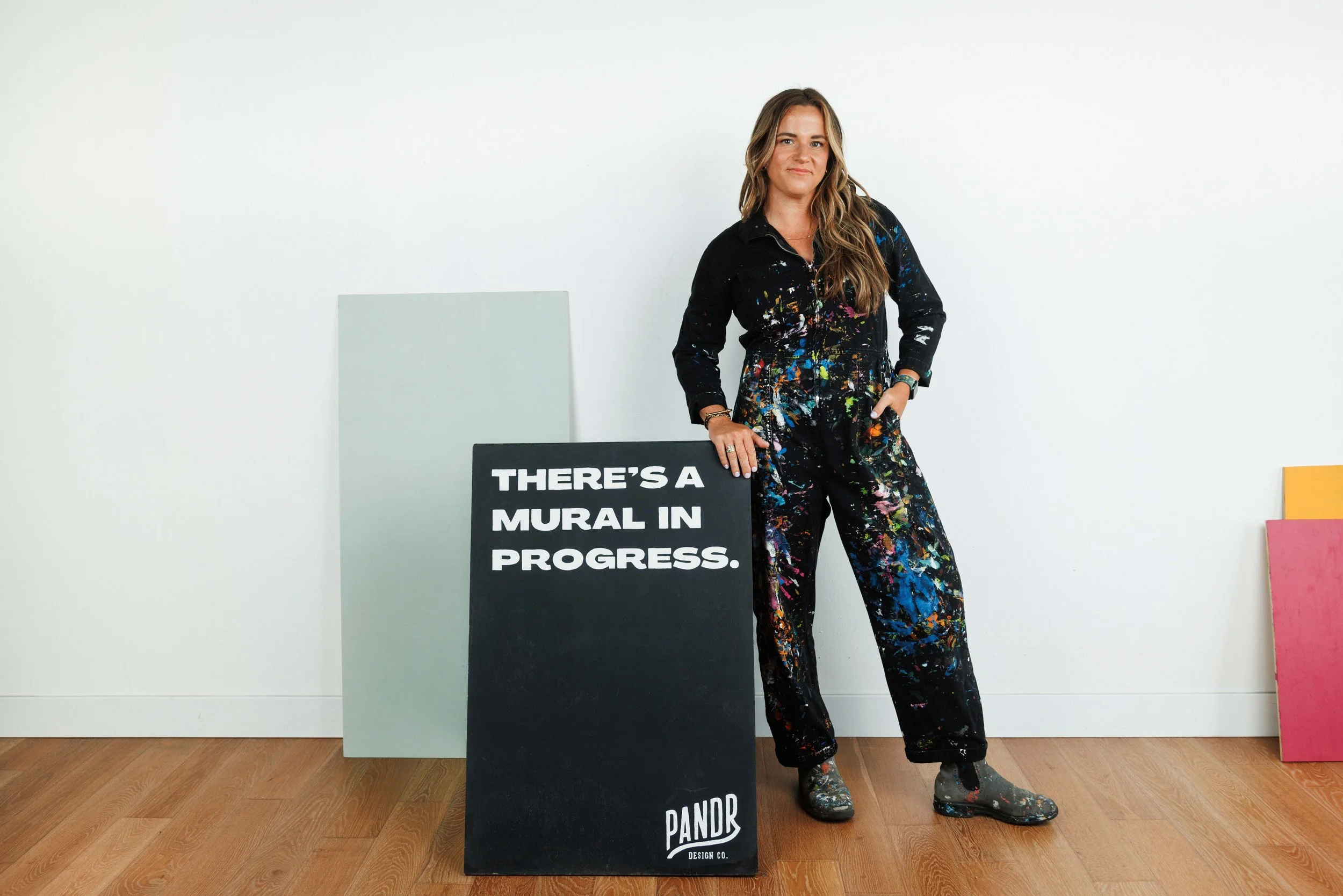 A San Diego mural artist in paint-splattered black jumpsuit stands beside a black sign that reads 'There's a mural in progress' in an art studio with blank canvases and a wooden floor.