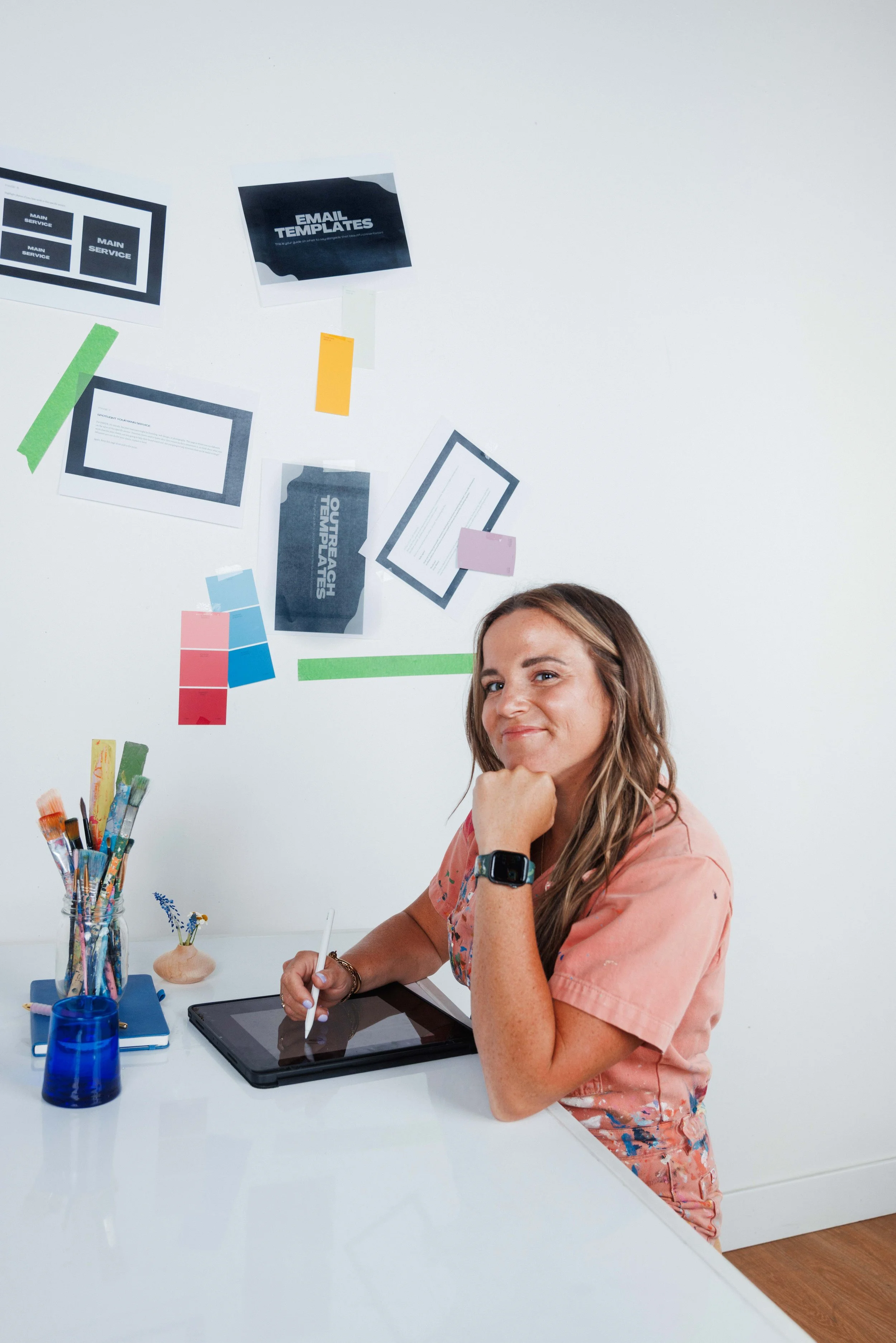 A woman with long brown hair sitting at a white desk, holding a stylus and using a graphics tablet, smiling and looking at the camera. On the desk are brushes, a blue container, a closed book, and a small vase with flowers. The wall behind her has papers, notes, and color swatches taped to it.