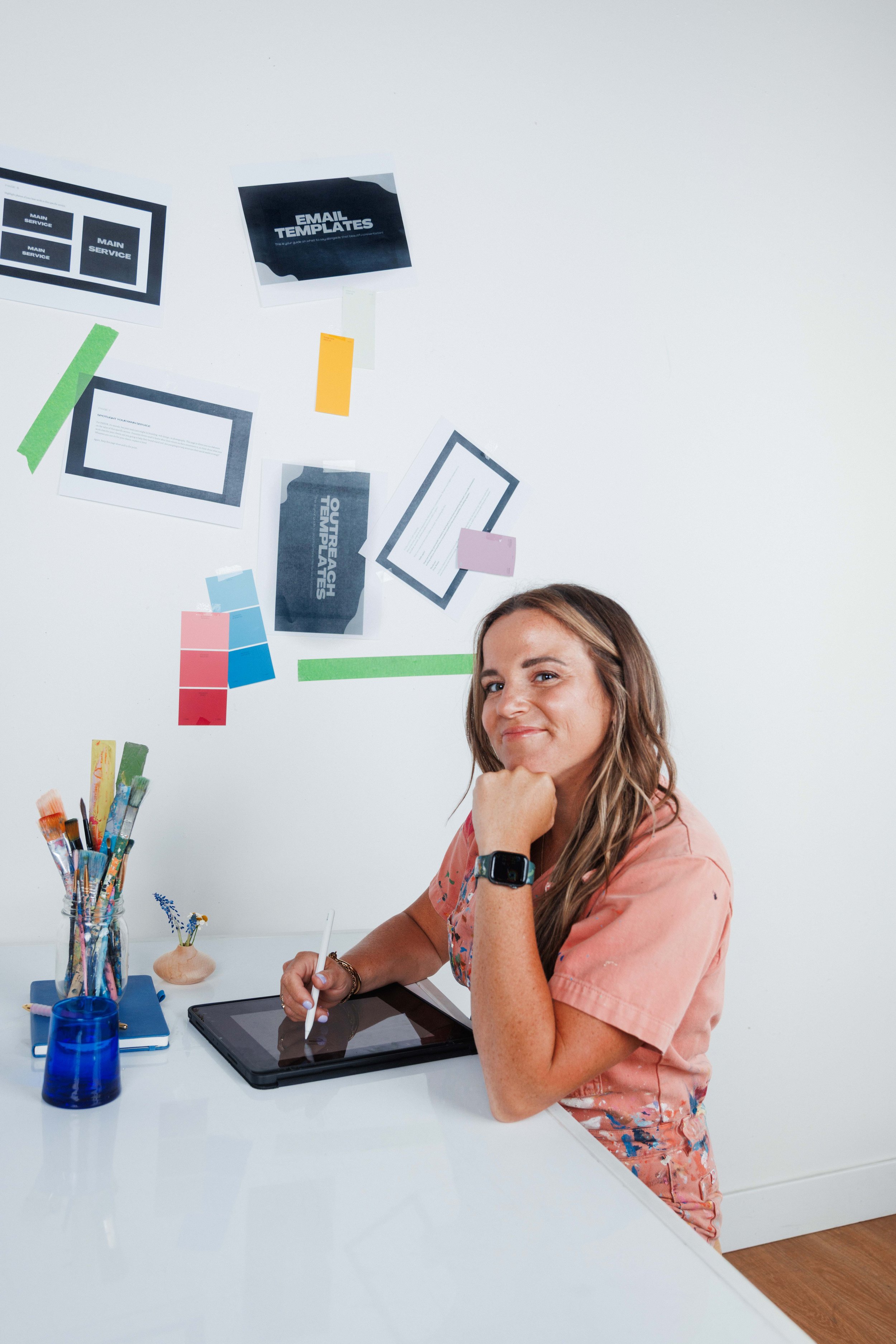 A woman with long brown hair sitting at a white desk, holding a stylus and using a graphics tablet, smiling and looking at the camera. On the desk are brushes, a blue container, a closed book, and a small vase with flowers. The wall behind her has papers, notes, and color swatches taped to it.