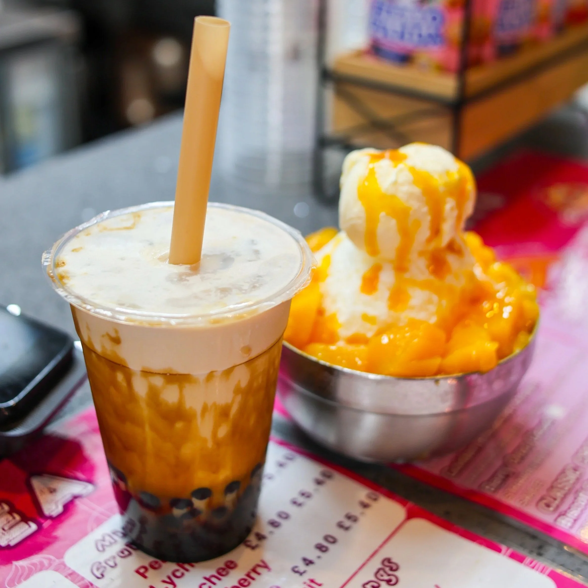 A tall plastic cup of bubble tea with a straw next to a bowl of shaved ice topped with mango chunks and mango syrup.