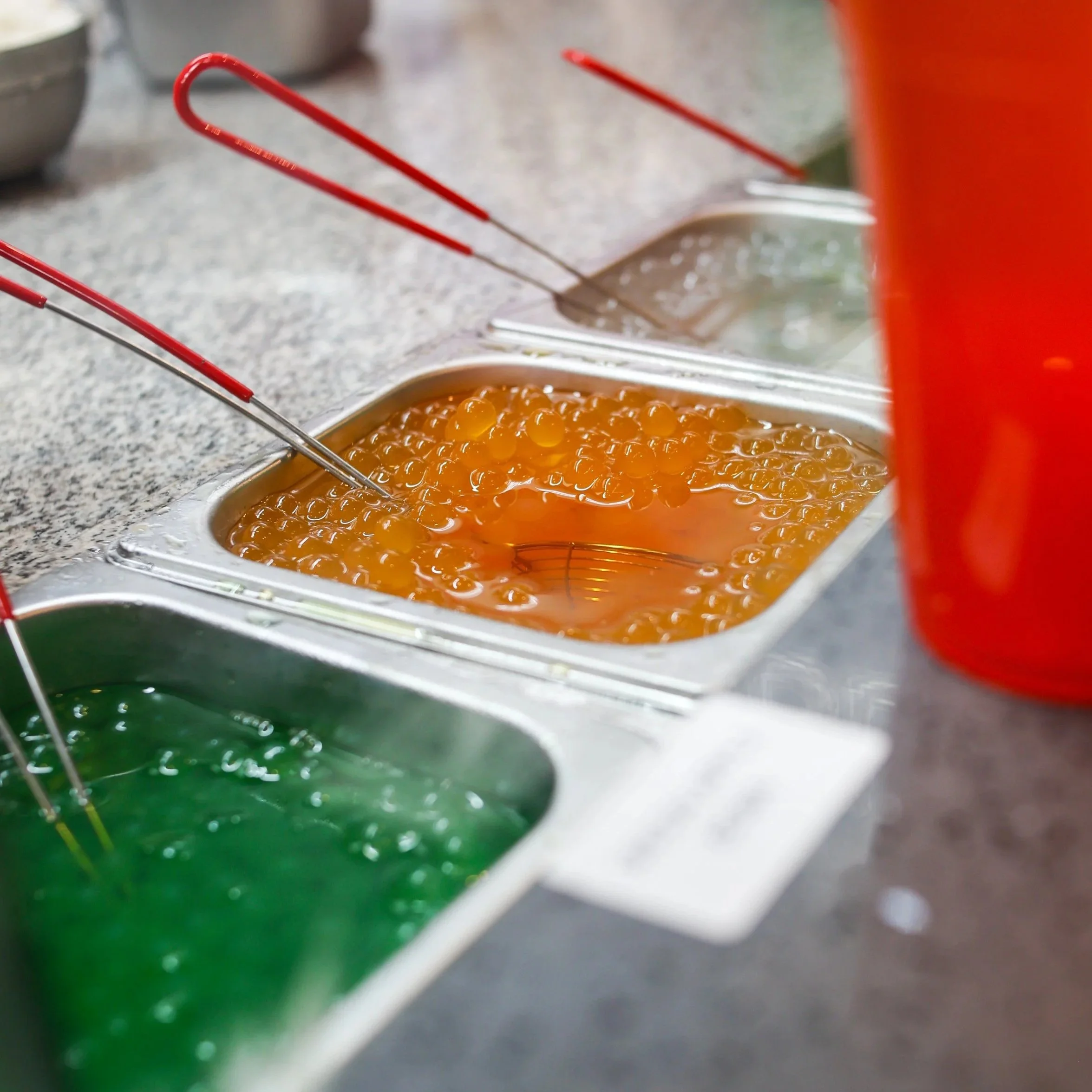 Container with orange and green bubble tea toppings and metal tongs for serving.